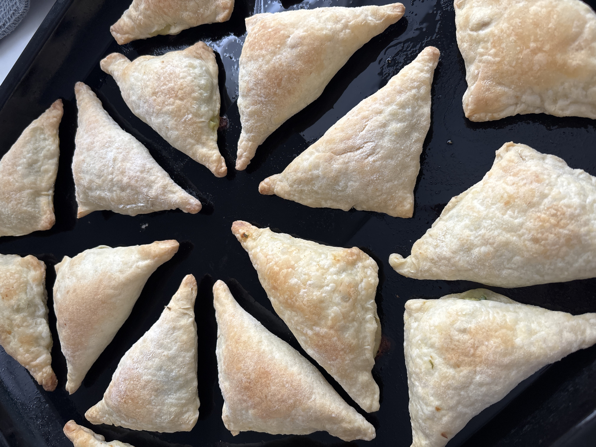 The image shows a black baking sheet containing approximately 15 triangle-shaped pastries. The pastries are golden brown and appear to be freshly baked. The pastries are arranged in a disorganized manner on the baking sheet. The baking sheet is resting on a light-colored surface. There is no indication of the city of Chaikovsky, Russia, or any people, actions or interactions.
