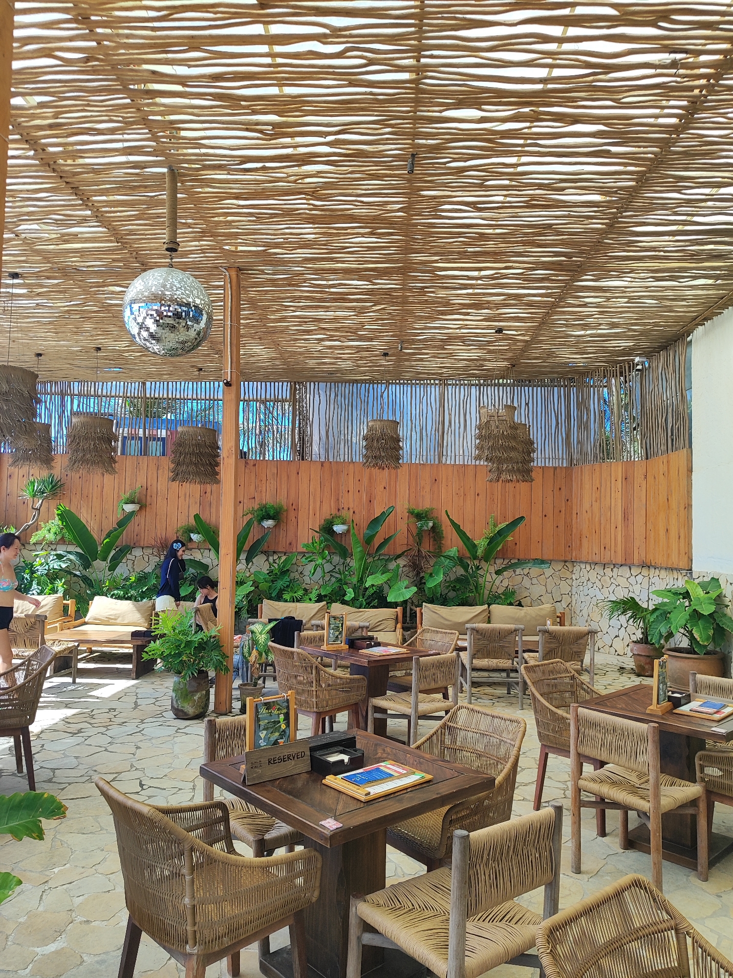 This image shows an outdoor seating area at a restaurant in Da Nang, Vietnam. The area is covered by a slatted bamboo roof and decorated with hanging lamps and a disco ball. There are several tables and chairs made of rattan, some with cushions. Plants are arranged throughout the seating area, creating a tropical atmosphere. In the background, there is a wooden fence and stone wall. Two women are visible in the image, one standing and one sitting, and a third person is partially visible in the background. One of the tables in the foreground has a "Reserved" sign on it.