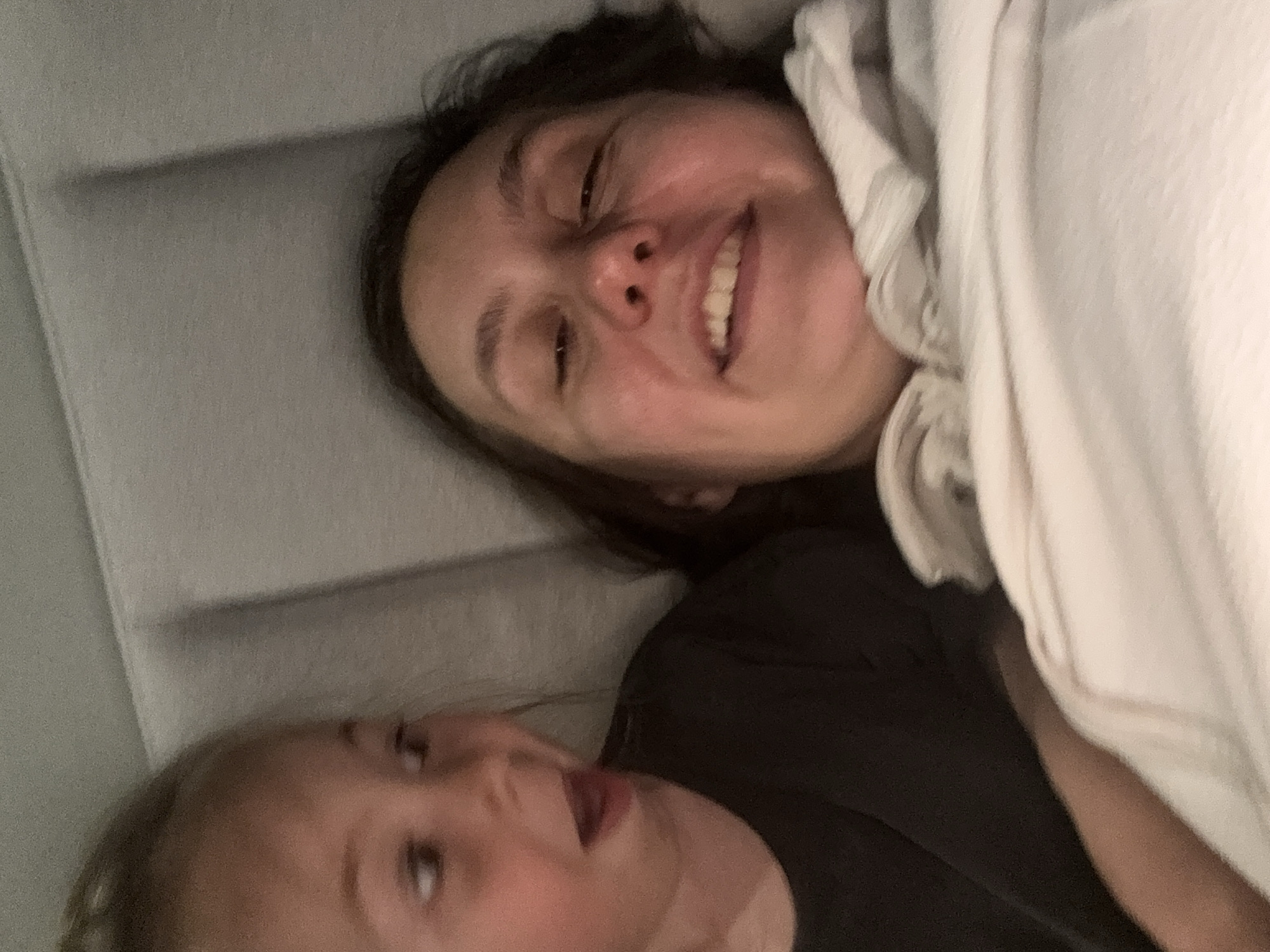A selfie-style image shows two people, seemingly a woman and a child, lying down. The woman is positioned towards the top of the frame, smiling with her eyes partially closed. She is partially covered with a white blanket or sheet. The child is located at the bottom of the frame, looking upwards. They are wearing a dark-colored shirt. The background appears to be a grey padded surface, possibly a headboard or wall. No information in the image allows any conclusion to be made regarding the location in Zelenogradsk, Russia.