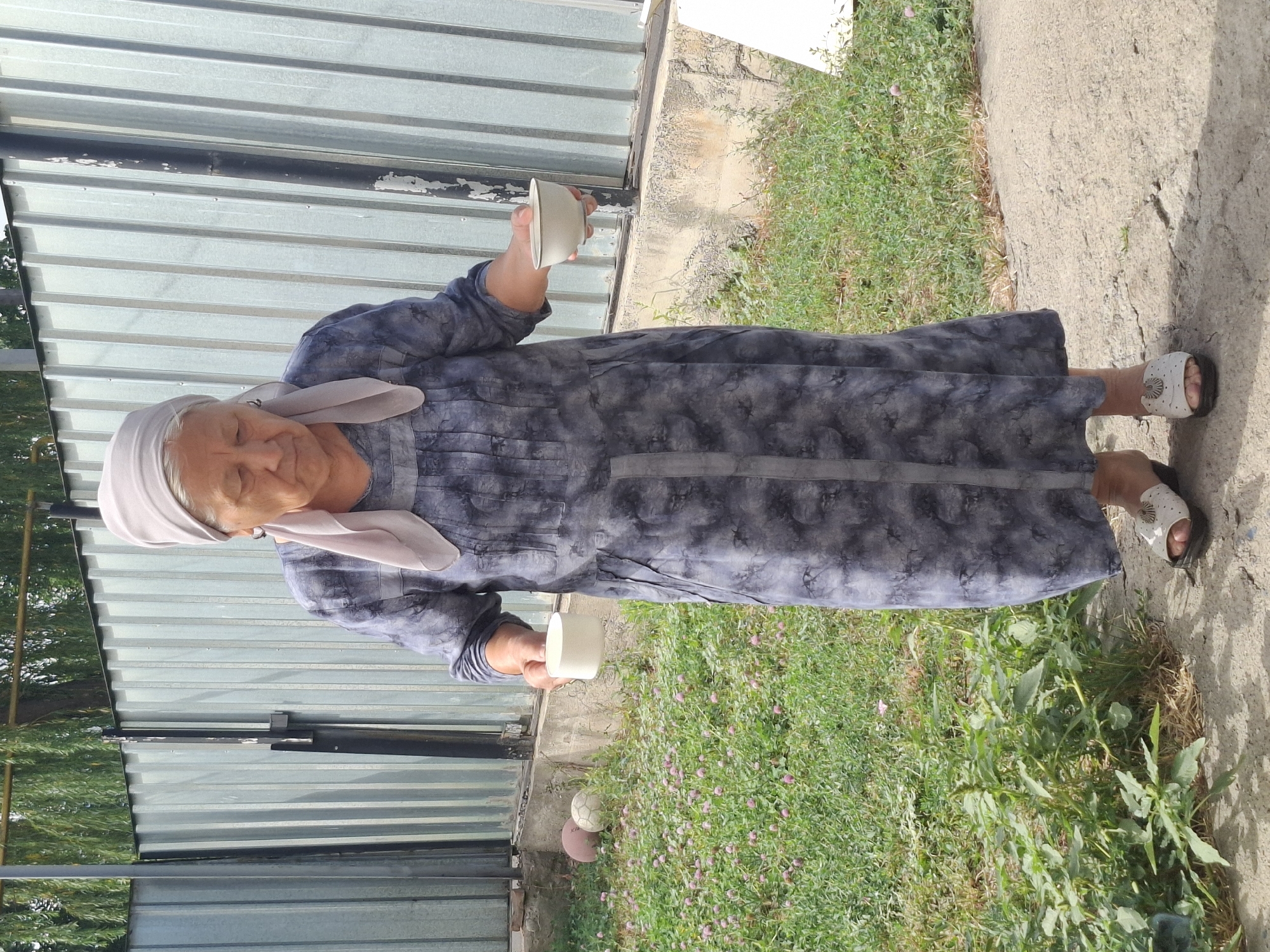 An elderly woman wearing a grey patterned dress and a light purple headscarf stands in a yard holding two white bowls. One bowl is held in her left hand, and the other in her right hand. Her gaze is directed downwards. The background features a corrugated metal fence, a concrete border, and a patch of green grass with small pink flowers. There are two balls visible on the grass to the left of the woman. The ground in front of her is paved with concrete. The overall scene suggests a residential outdoor setting, possibly a backyard.