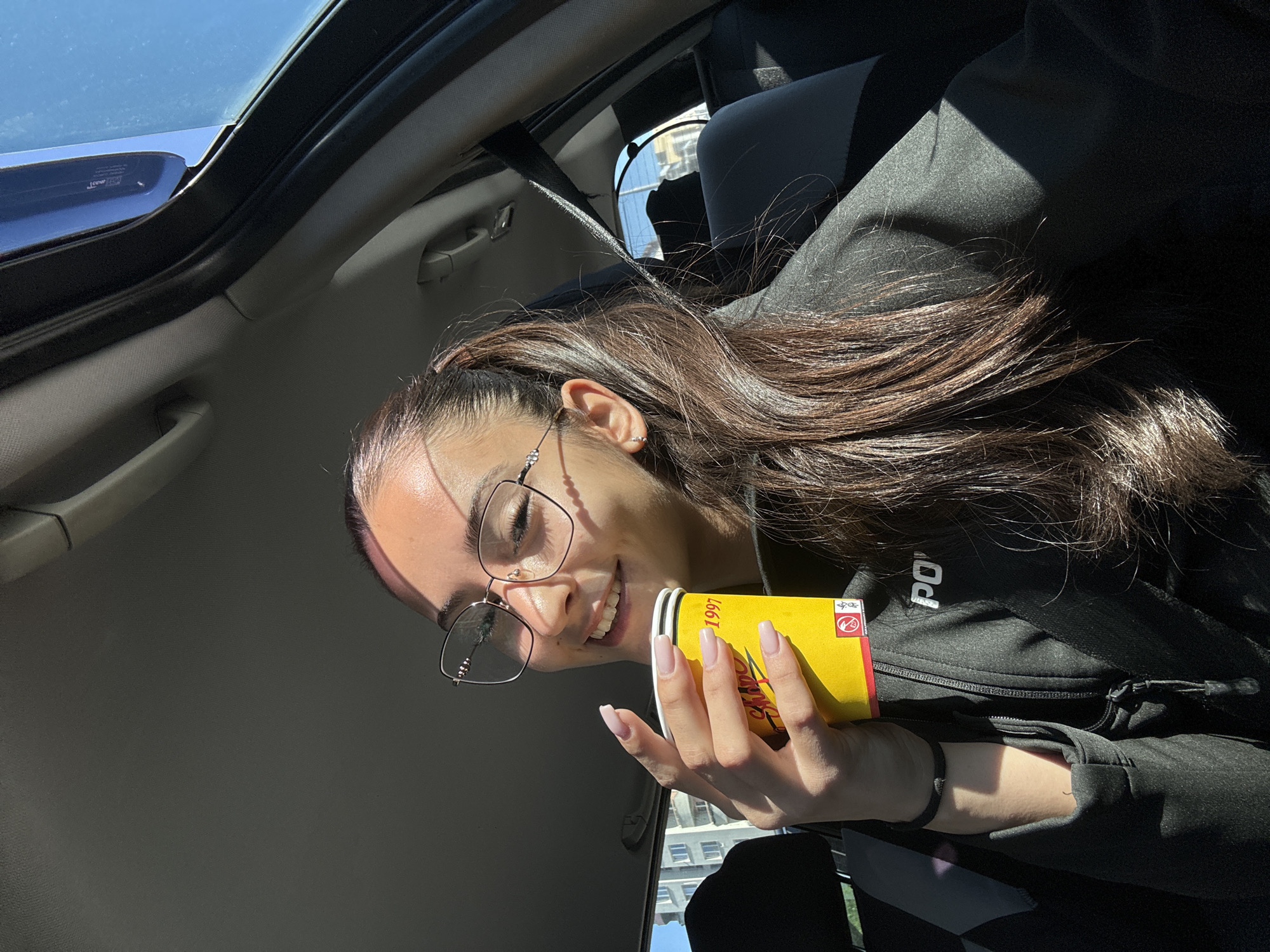 The image shows a young woman inside a car, likely a passenger, with a sunroof open above her. She is holding a yellow drink container with text and what appears to be a "1997" label. She is wearing glasses and a dark jacket or top. Her long brown hair is partially pulled back. The background visible through the car window includes a building exterior, suggesting an urban setting. The location is likely Sofia, Bulgaria.
