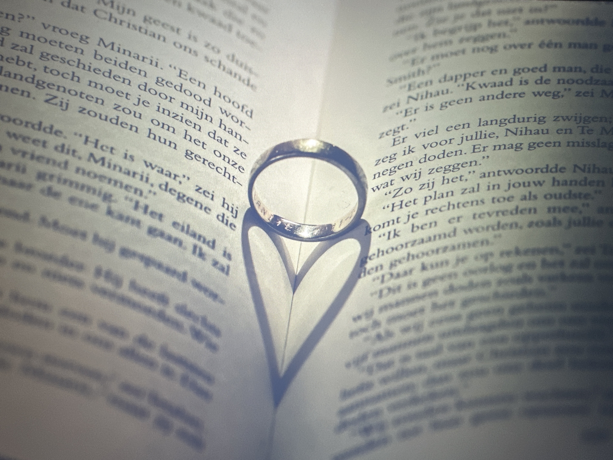 The image shows a close-up of an open book with text in Dutch. A gold wedding ring is placed on the open pages of the book. The ring casts a shadow in the shape of a heart on the right page of the book. The book appears to be lying flat on a surface, possibly a table. The lighting suggests the photo was taken indoors, possibly with artificial light. There are no people or any other objects visible in the image, and no information is provided about the location in the city of Bryansk, Russia.