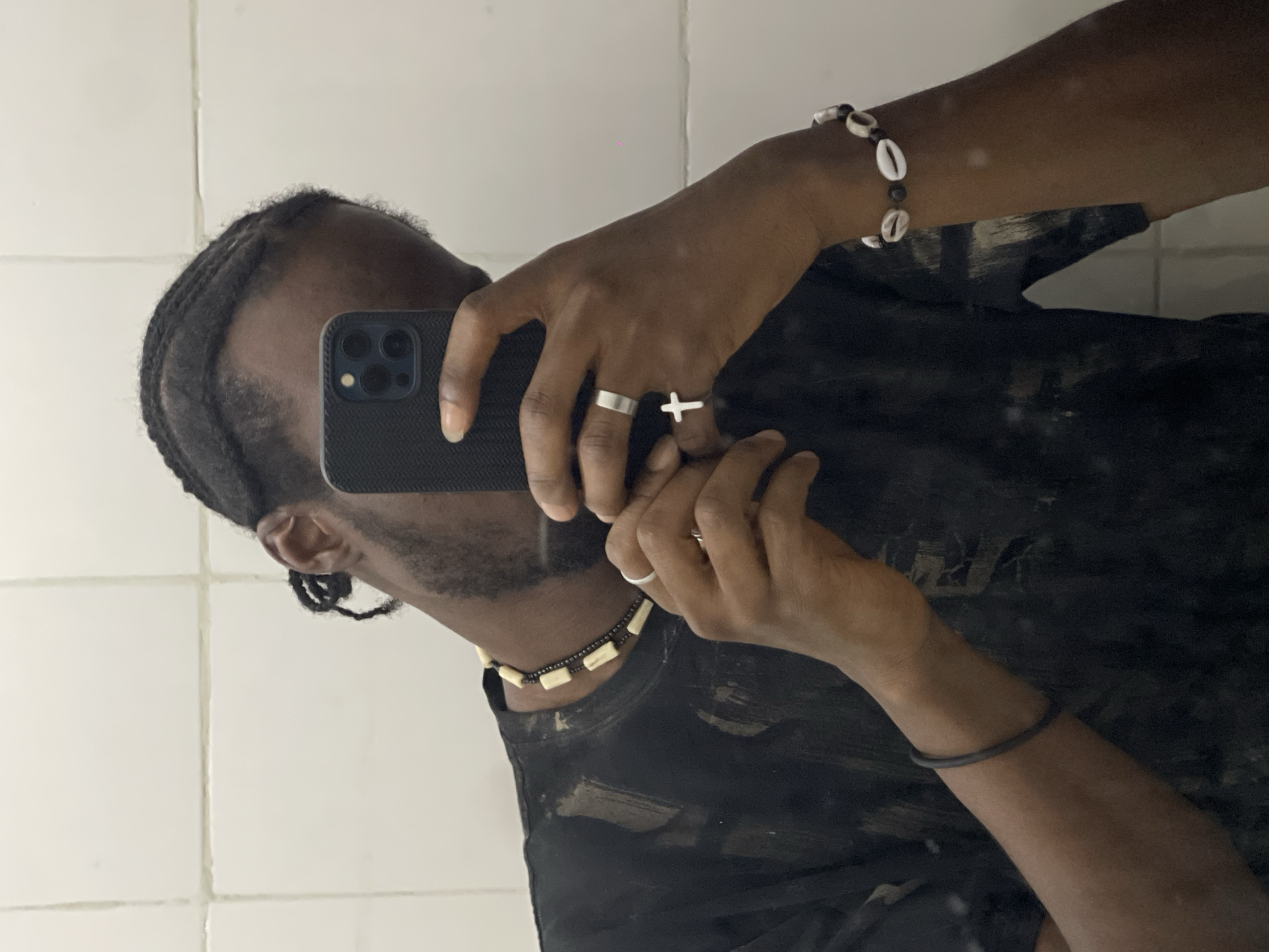 A person is taking a selfie in front of a white tiled wall, presumably a bathroom. The person has braided hair and is wearing a black t-shirt. Visible accessories include a necklace with light-colored rectangular pieces, a bracelet with seashells, a thin bracelet on the left wrist, and two rings on the right hand—one a plain band and the other a cross. The person is holding a black smartphone up to their face. The image does not provide specific information to confirm that it was taken in Jos, Nigeria.