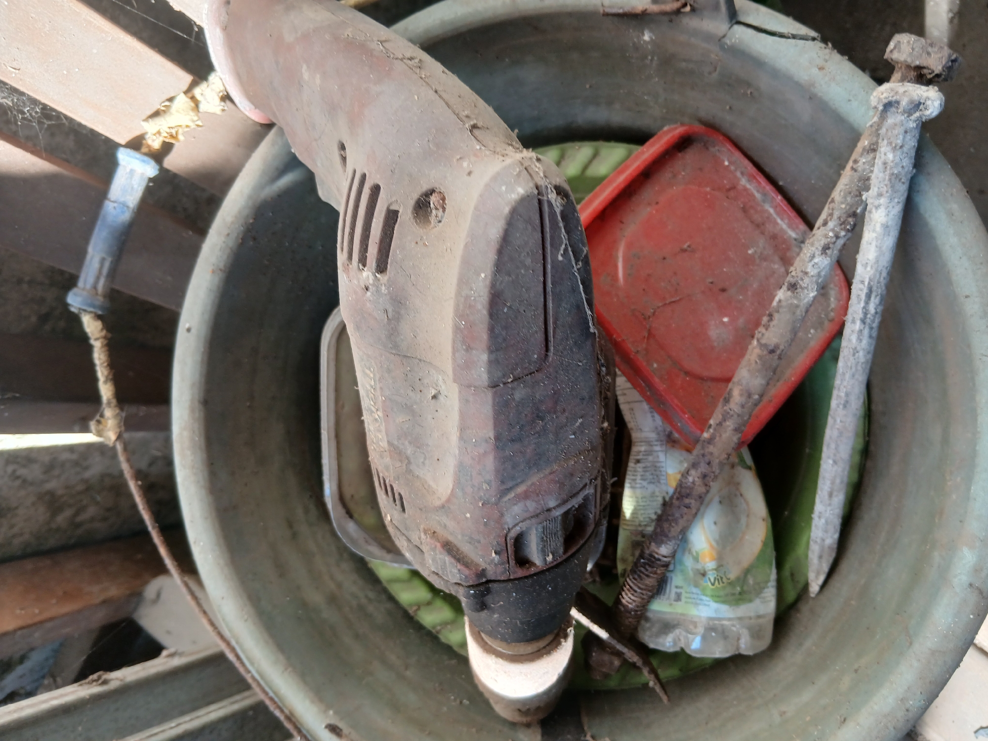 The image depicts a collection of dusty tools and debris inside a large, green plastic basin. Dominating the center is a red and black power drill, covered in a layer of dust and cobwebs. To the left of the drill, a clear plastic bottle with a green label is visible. In front of the bottle, a metal rod with a pointed end and another rusted metal object with a hexagonal nut are positioned. A red, square-shaped plastic lid is situated behind the drill. The basin itself appears worn and dirty. In the background, wooden planks and a rope are visible, suggesting an outdoor or storage area. The overall impression is one of disarray and neglect. The scene is situated in the city of Tenggarang, Indonesia.