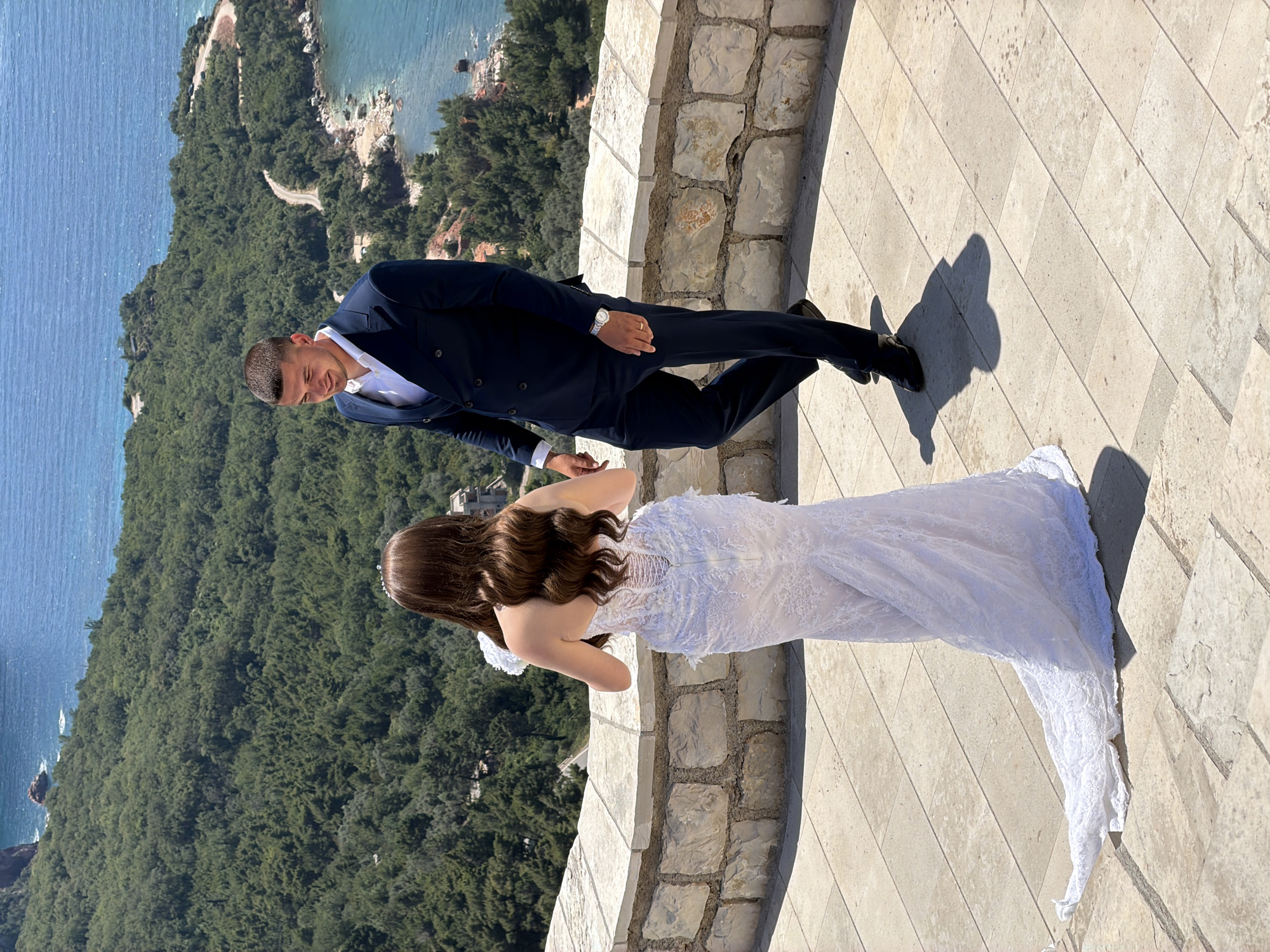 An over-the-shoulder shot depicts a bridal couple on a stone terrace with a backdrop of lush green foliage and the azure Adriatic Sea, suggesting a location in or near Sveti Stefan, Montenegro. The bride, facing away from the camera, wears an elaborate white lace wedding gown with a long train that trails on the stone paving. Her long, dark, wavy hair is visible. She holds a white bouquet. The groom, facing the bride, is dressed in a dark navy double-breasted suit with a white dress shirt. He is looking down and to the side, with a slight smile. The terrace is bordered by a low stone wall. A winding road is visible through the trees in the distance, leading to a coastline with rocky outcrops. The sun casts distinct shadows on the terrace.