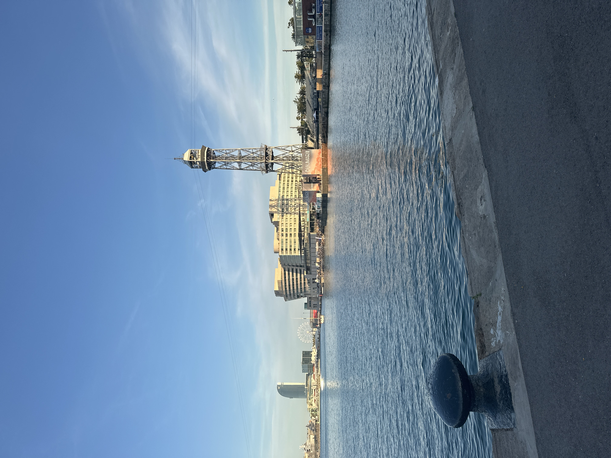 The image depicts a waterfront scene in Barcelona, Spain. In the foreground, a dark, bollard sits on a concrete pier beside an asphalt path. The water of the harbor is calm, with ripples reflecting the blue sky. Across the water, a tall, industrial-style tower with a cage-like structure stands prominently. Behind the tower, a long, multi-story building with a light-colored facade and many windows stretches along the waterfront. Further in the background, other buildings, including a tall, modern skyscraper with a "W" logo, a Ferris wheel, and boats, are visible. The sky is a clear blue with a few wispy clouds, suggesting it is daytime. The overall atmosphere is one of a bustling port city with architectural landmarks.