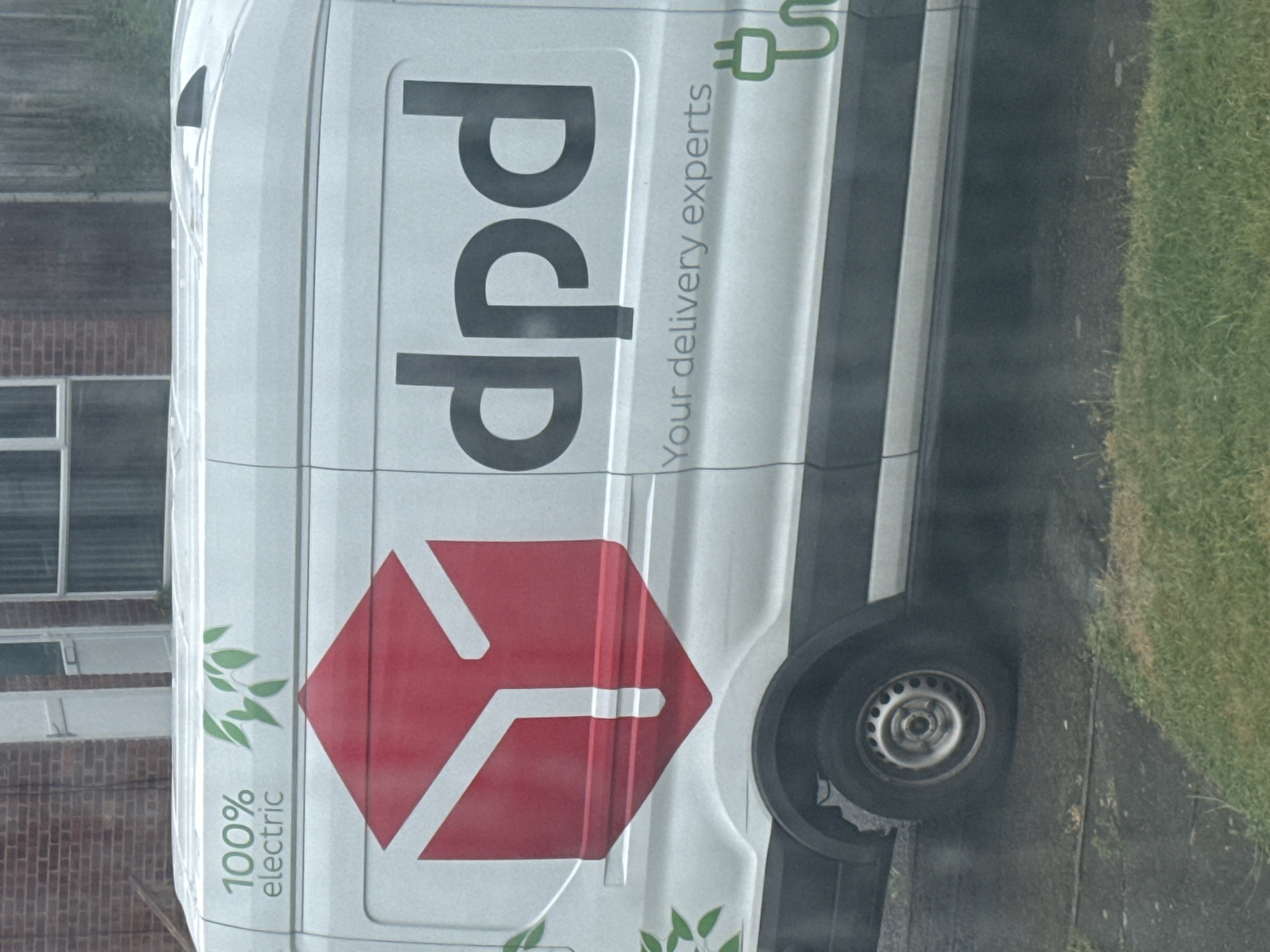 A white DPD delivery van is depicted in a daytime outdoor scene, possibly during or after rain, given the damp appearance of surfaces and slight haziness. The van is parked on a paved area, with a strip of green grass visible to its right. In the background, part of a brick building with several windows is visible.
The van's side is prominently branded with the DPD logo, featuring large black letters spelling "dpd". Below this, vertically oriented text reads "Your delivery experts". To the left, also oriented vertically, is the text "100% electric", accompanied by green leaf motifs and a green electric plug icon, emphasizing the vehicle's eco-friendly nature. A large, red hexagonal DPD logo with white geometric lines is positioned towards the rear side of the van. The van's front wheel is visible in the lower right portion of the image. No people are visible, and the scene suggests the van is stationary, likely parked.