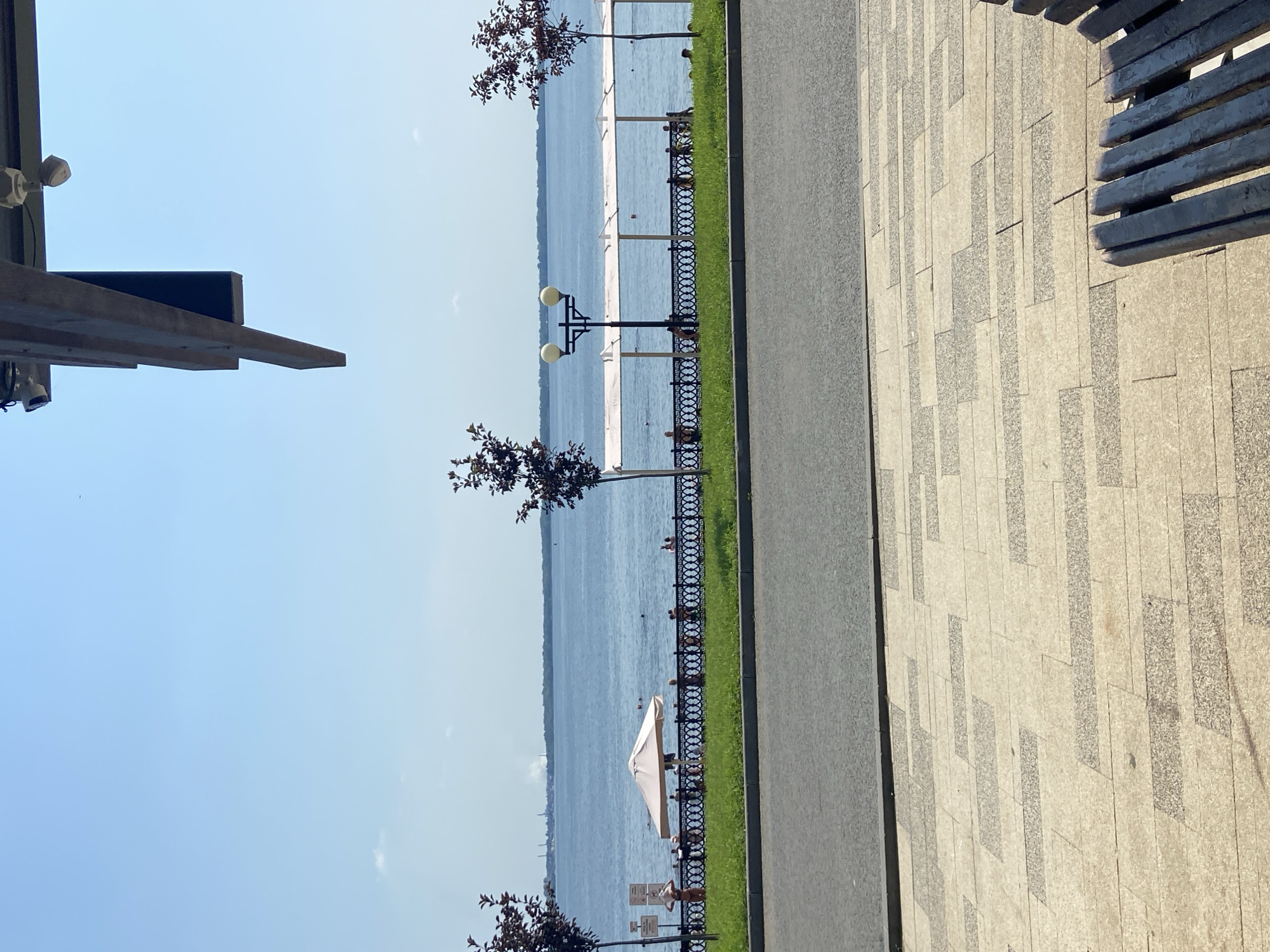 The image depicts a sunny day at a waterfront promenade in Saratov, Russia. In the foreground, a paved walkway with a patterned design leads to a dark wooden bench partially visible on the right. Behind the walkway is a strip of green grass, followed by a black decorative metal fence. Beyond the fence, a sandy beach area is visible, with several people sunbathing and a few individuals in the water. An open umbrella provides shade on the beach.
Further back, a wide blue body of water stretches to the horizon. On the opposite shore, a distant cityscape is visible with several industrial structures emitting plumes of smoke. Along the waterfront, a modern structure with white canopies is present, offering shaded areas. A lamppost with two globes stands near the water. Two small trees with dark foliage are positioned on either side of the promenade, framing the view of the water. The sky is clear and blue, with a few wispy clouds. Overhanging the top portion of the image is a dark wooden structure, possibly part of a building or canopy, with attached security cameras and lights. The overall scene conveys a sense of a recreational area on a pleasant summer day.
