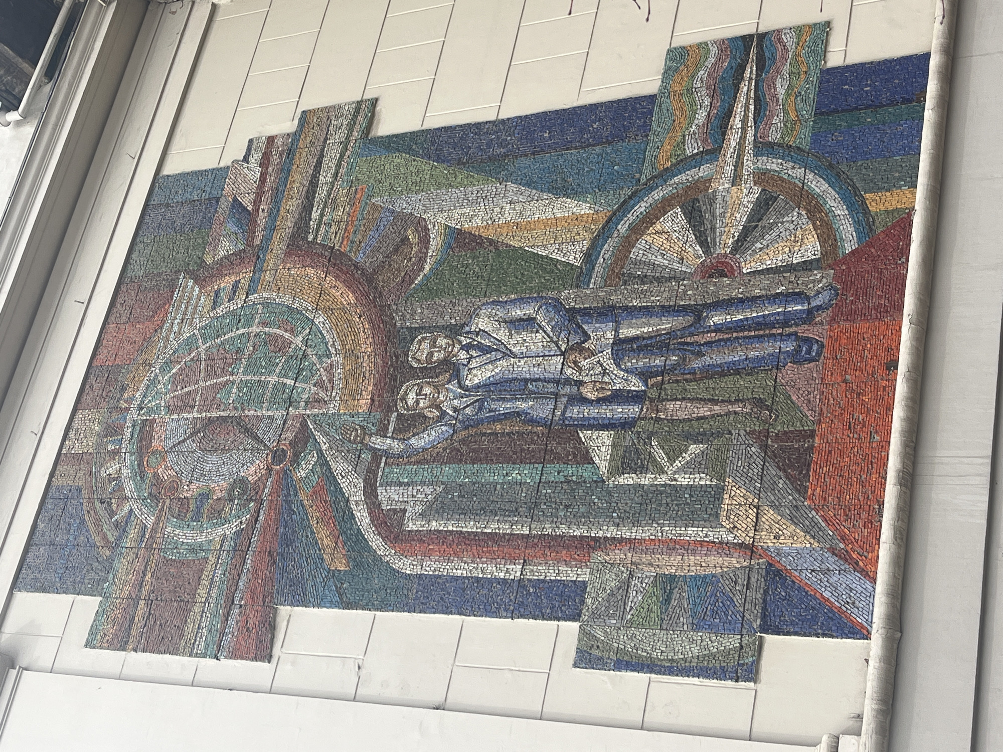 A low-angle, full shot captures a mosaic mural. The mural features two figures dressed in blue suits lying horizontally across the image center. The figures appear to be superimposed on a backdrop with stylized representations of machinery, including wheels and gears. The color palette is diverse, incorporating blues, greens, browns, reds, and whites in small, mosaic tiles. The mural is positioned against a white tiled wall. The top left corner of the image shows the underside of a ceiling with visible pipes and structural elements. A vertical column with visible joints is on the right.
The image does not contain the specified location details.