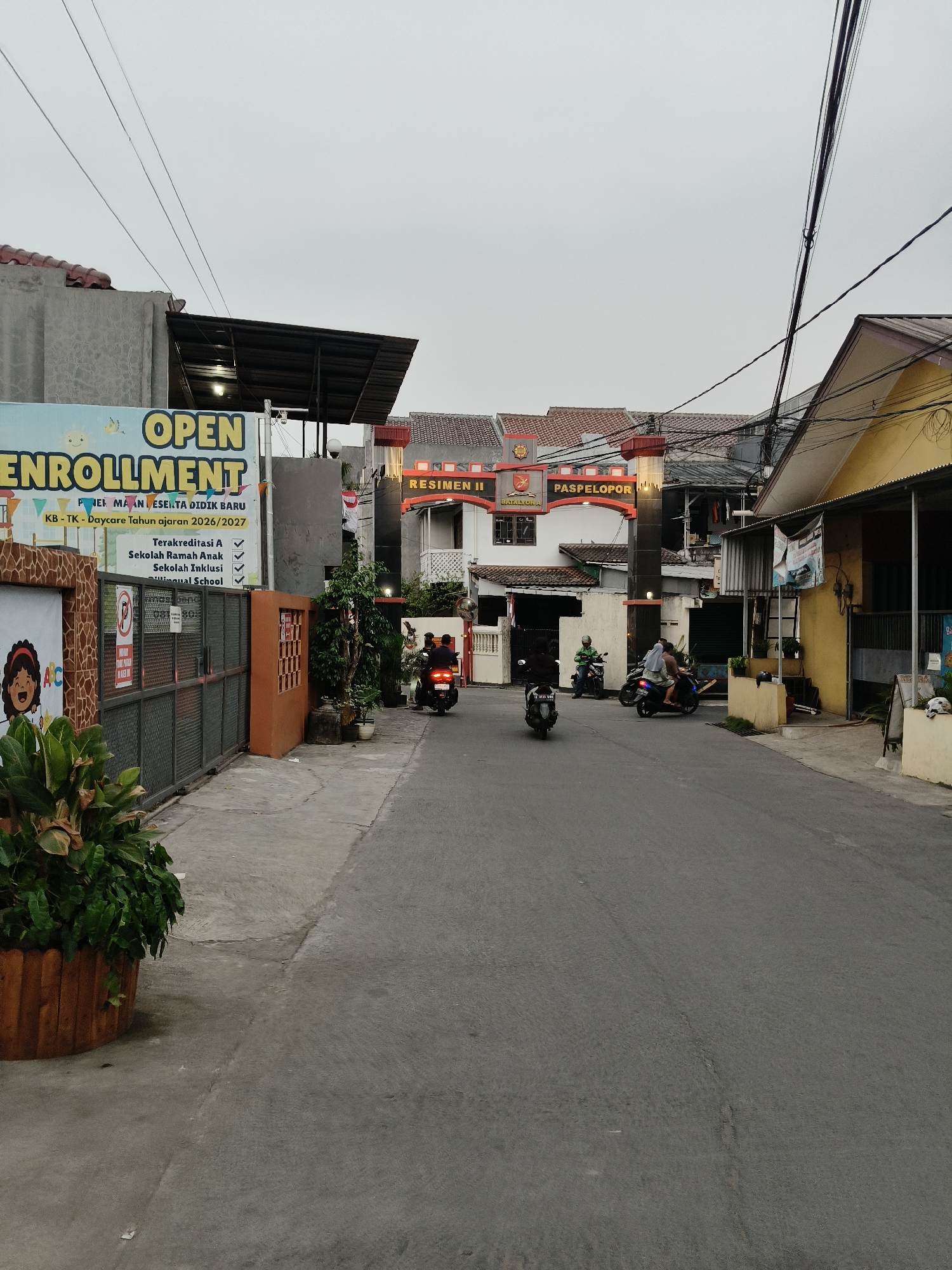 The image depicts a street scene in South Tangerang, Indonesia, likely within a residential or school area, captured on an overcast day. Dominating the left side of the frame is a large, blue and yellow sign advertising "OPEN ENROLLMENT" for "KB-TK - Daycare Tahun ajaran 2026/2027," indicating a kindergarten and daycare facility. Below this, text lists accreditations and school types: "Terakreditasi A," "Sekolah Ramah Anak," "Sekolah Inklusi," and "Bilingual School." Adjacent to the sign is a metal gate and a wall decorated with a stylized image of a child. A large potted plant with dense green foliage is positioned in the foreground on the left.
The street itself is paved and appears to be a two-lane road. Several motorcycles are visible on the street. In the mid-ground, a white archway with "RESIMEN II PASPELOPOR" inscribed on it serves as an entrance to a complex. This archway is flanked by buildings with red-tiled roofs. On the right side of the street, a yellow building with a covered awning and various signs is visible. Power lines crisscross the sky. The overall impression is a quiet urban street with a focus on an educational institution.