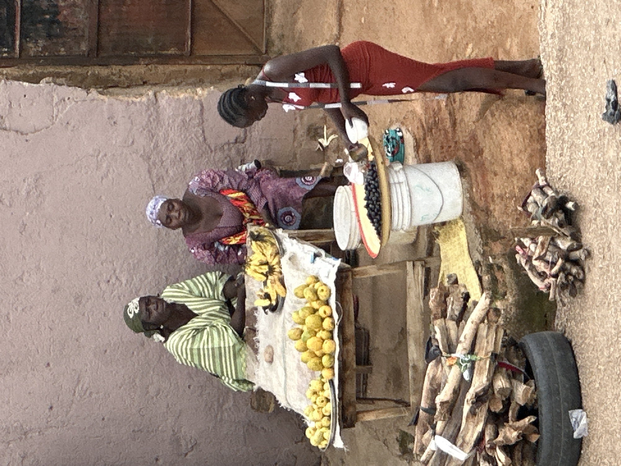 In Jos, Nigeria, two women sit behind a table laden with fruits, likely bananas and citrus, while another woman stands to the right, attending to a sale. The woman on the left wears a green and white striped robe and a green headwrap. The woman in the middle is dressed in a purple and pink patterned dress. The woman on the right, dressed in a red dress, is pouring something from a small white cup into a larger white bowl. A pile of firewood is visible in the lower left corner of the image, next to a tire. The ground is made of dirt and gravel. The background consists of a textured wall and a wooden door.