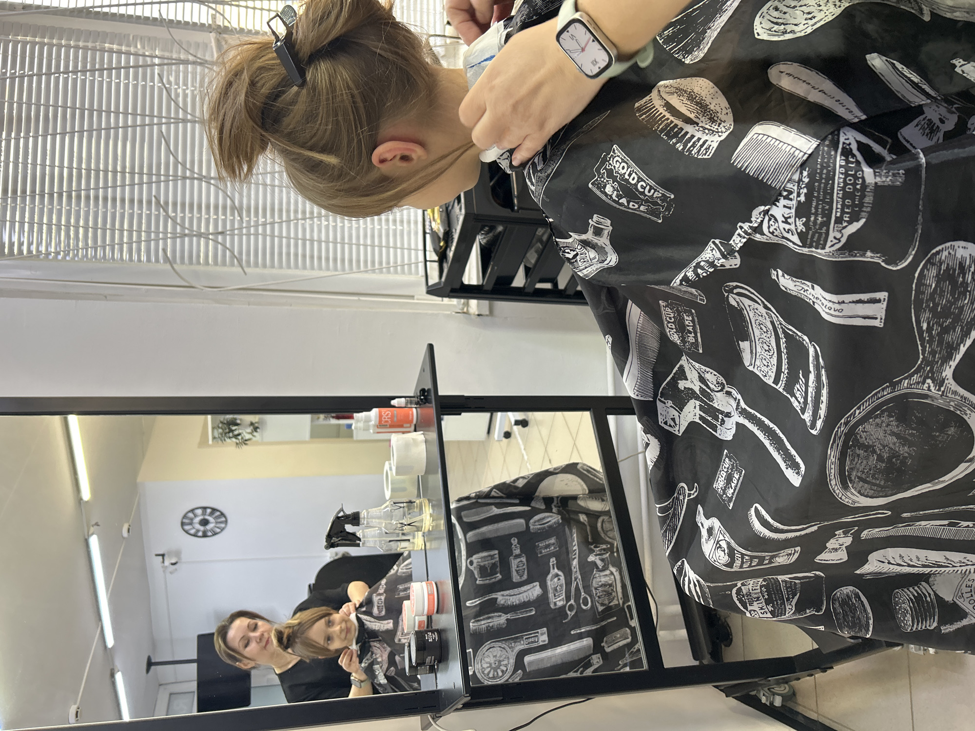 A woman is getting a haircut in a hair salon. The woman's hair is in a bun, and a clip is holding some of her hair in place. She is wearing a cape that is black with white drawings of barber tools. The barber is wearing a light green Apple Watch on her wrist. The salon has white walls and bright lighting. There is a mirror that reflects the woman getting her haircut and the barber.
There is no evidence that this image was taken in Moscow, Russia.