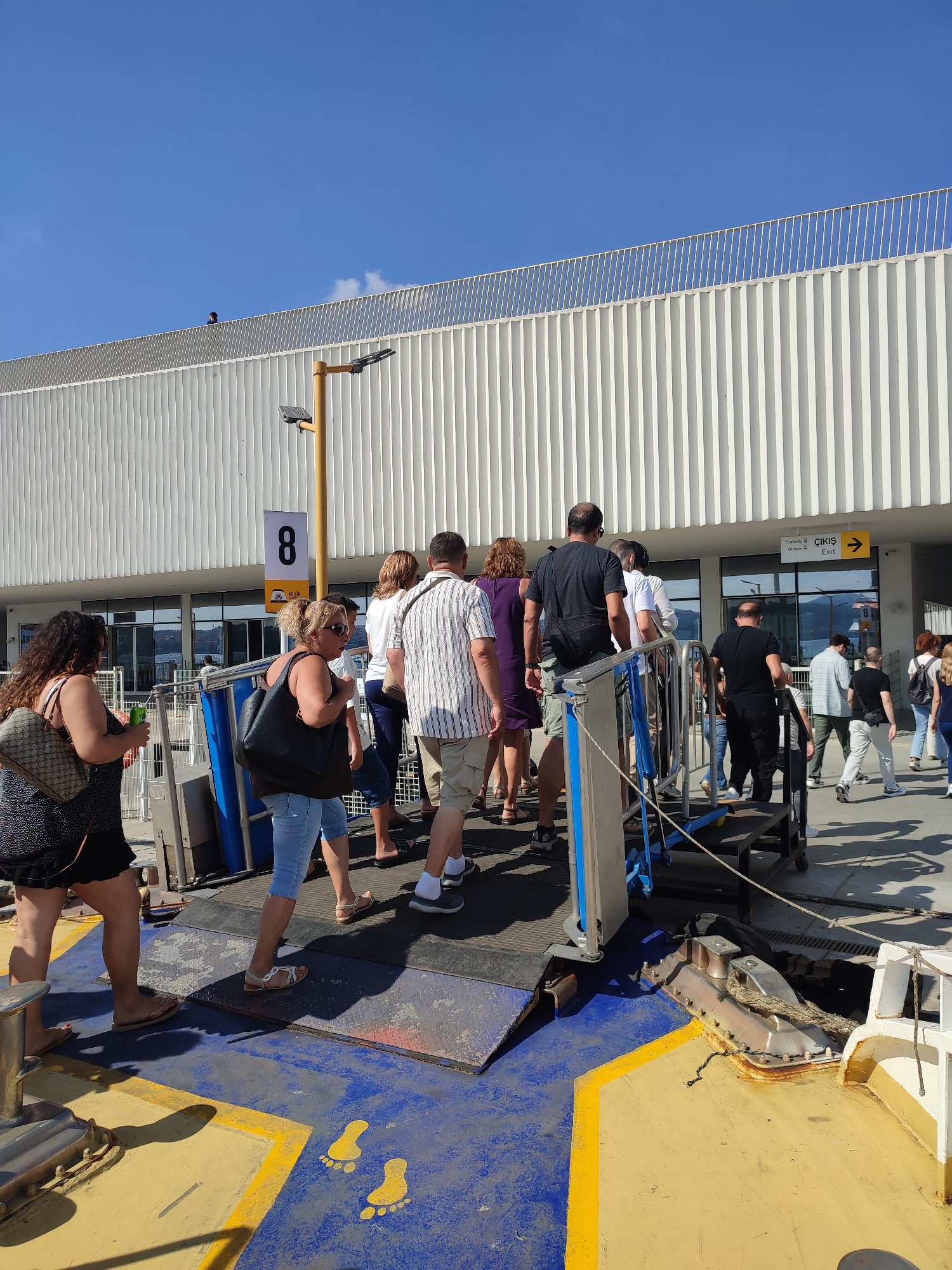 The image depicts a group of people disembarking from a ferry onto a ramp. The ramp is painted with yellow and blue markings, including yellow footprint graphics. The ferry appears to be docked at a terminal building with a modern, white, ribbed facade. A sign with the number "8" is visible on a yellow pole, along with directional signage indicating "Tramvay" and "Otobüs" (bus) with an arrow pointing right, and "ÇIKIŞ" (Exit). Several individuals are walking up the ramp, with some carrying bags. In the background, the sky is clear blue. The context suggests this is a transportation hub, likely a ferry terminal in a coastal city. Considering the signage with Turkish words, this location is identified as the Beyoğlu, Turkey ferry terminal.