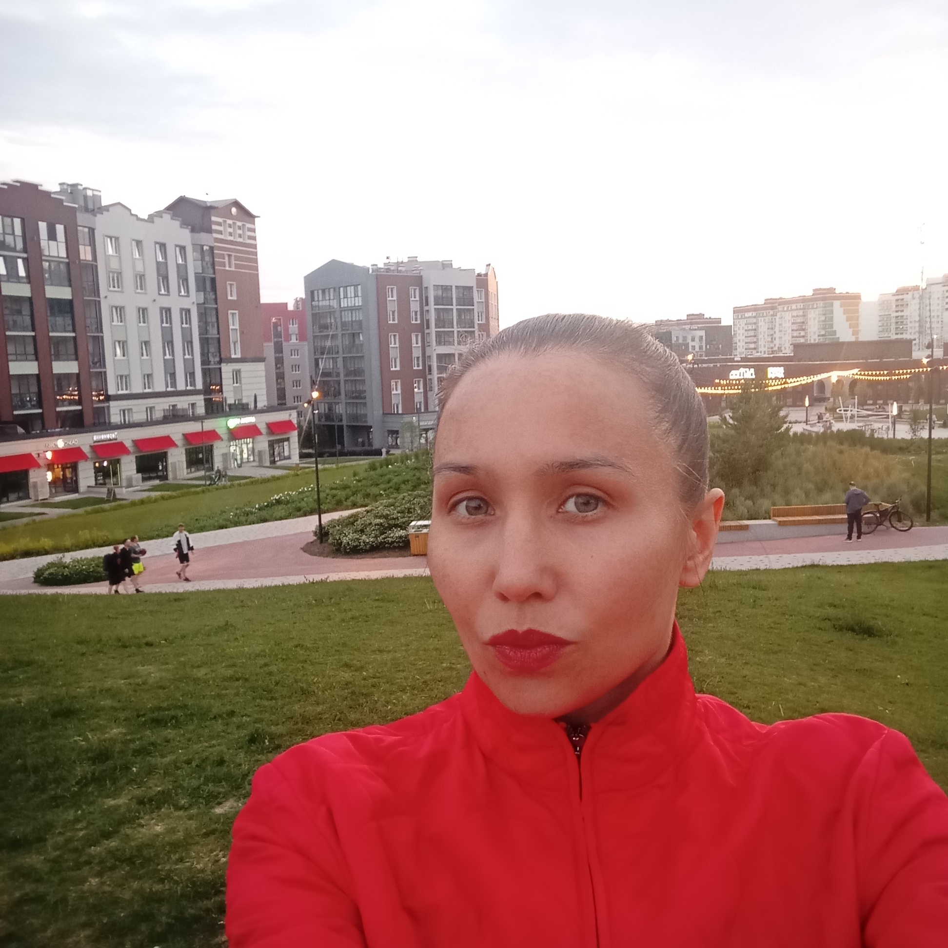 The image is a selfie taken outdoors in what appears to be a modern urban park or residential area. The background features several multi-story buildings with modern architecture, characterized by clean lines, multiple windows, and balconies. Some buildings have red awnings over storefronts on the ground floor. A paved walkway curves through a grassy area, and in the distance, there are benches and streetlights. The sky is overcast and bright, suggesting either early morning or late afternoon.
In the foreground, a woman is taking the selfie. She has her hair pulled back and is wearing a bright red jacket with a zipper. Her facial features are clearly visible, including her eyes, nose, and lips, which are colored with red lipstick. To the left of the woman, in the mid-ground, three people are walking on the paved path. Further to the right, a person is standing next to a bicycle near a bench. The overall scene conveys a sense of an active or recreational urban environment.
The location in the city of Pervomaysky, Russia, is confirmed by the presence of a sign that reads "astra Rr..." which is a known business in Pervomaysky.