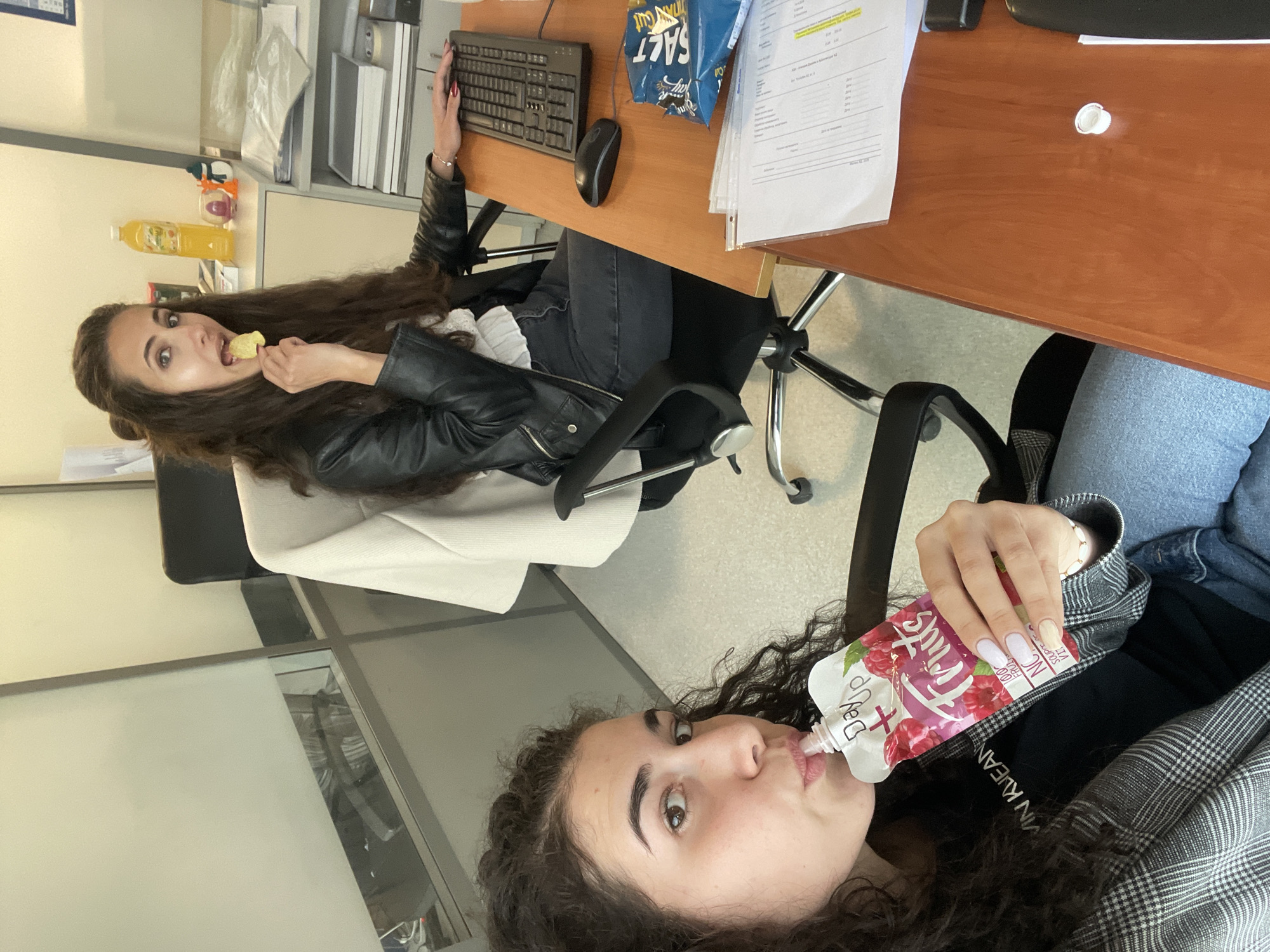 Two women are in an office setting. The woman in the upper part of the image is seated at a desk, wearing a black leather jacket and eating a chip. The desk has a computer keyboard, mouse, a bag of chips, and documents. The woman in the lower part of the image is drinking from a fruit puree pouch. Her clothing includes a plaid jacket. The location is indicated by the text "VIN KVEAN" on her clothing. The location in Sofia, Bulgaria is not deducible.