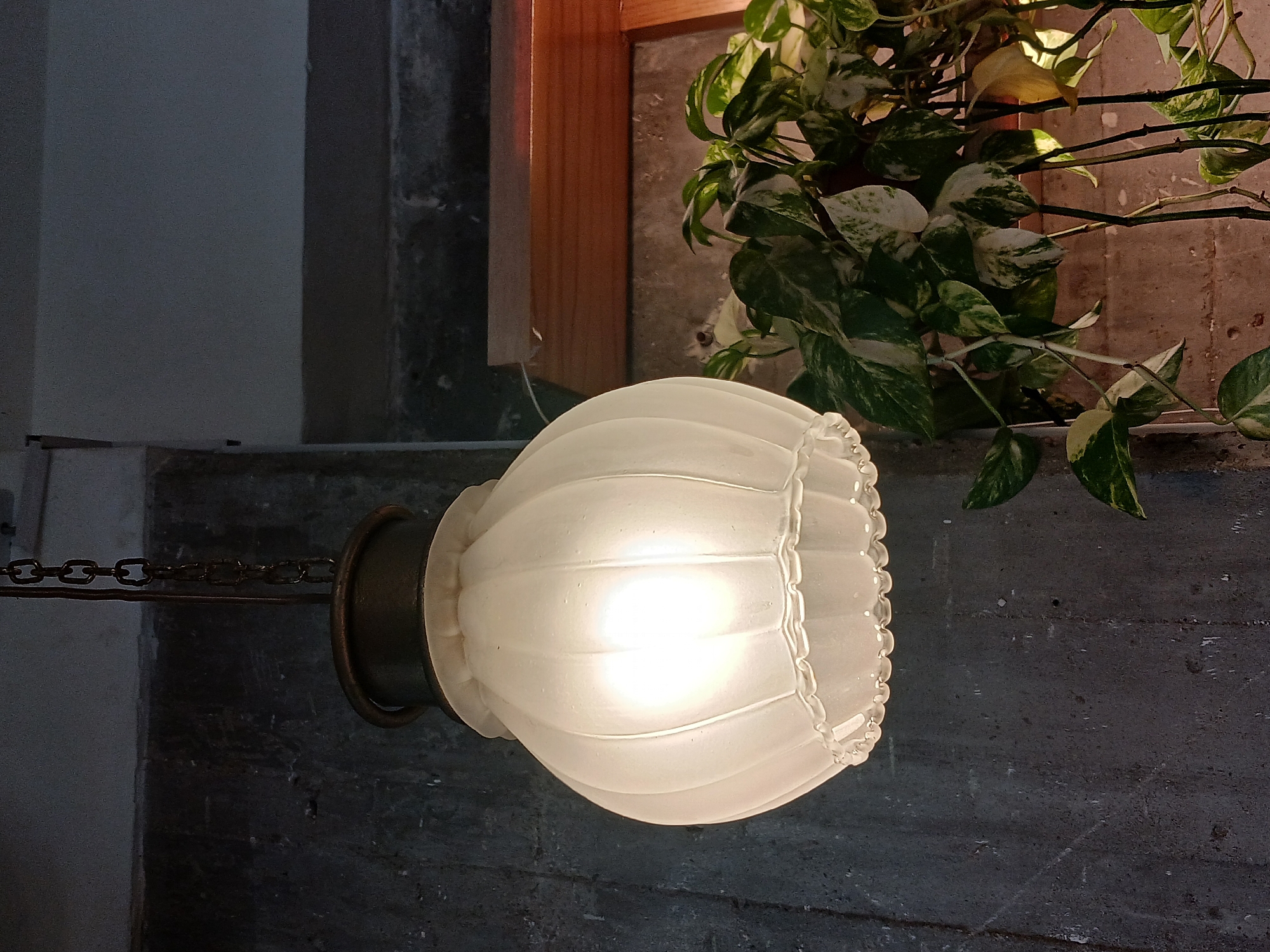The image shows a lit frosted glass lamp hanging from a chain, with a potted plant in the background. The lamp is round and has a ruffled edge. The plant has green and white variegated leaves. The background appears to be a wall with a wooden frame. The specific location in Buenos Aires, Argentina, is not discernible from the image.