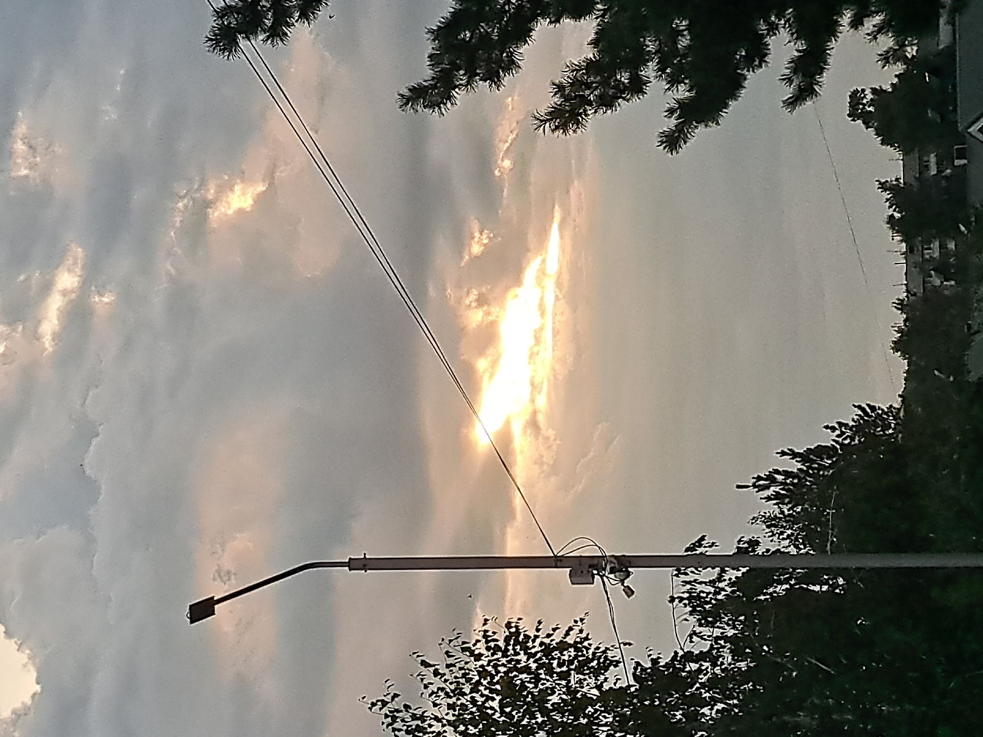 The image shows a street lamp in the foreground, with wires extending across the frame. Behind the lamp, a dramatic sky is visible, with the sun breaking through dark clouds, creating a bright, illuminated patch. The lower portion of the image features dark silhouettes of trees and some buildings in the distance. The scene appears to be an outdoor urban environment, possibly during dusk or a cloudy day. The location in Irkutsk, Russia is not explicitly indicated by any visible landmarks or text within the image.