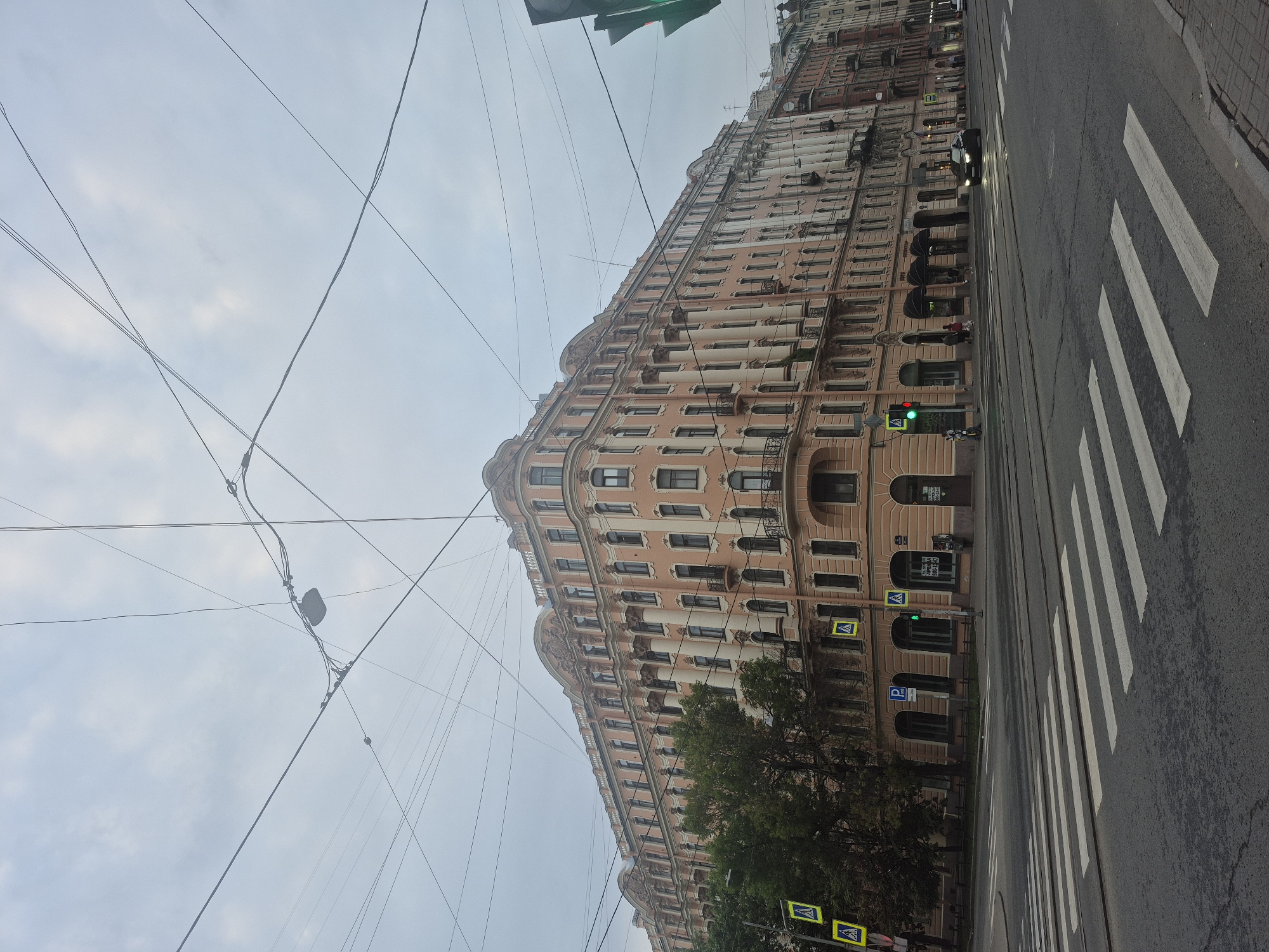 The image depicts a street scene in Saint Petersburg, Russia, characterized by an overcast sky. A long, ornate, pale pink building with many windows lines the left side of the frame. The building exhibits classical architectural elements, including decorative columns and intricate detailing. Overhead, a complex network of electrical wires crisscrosses the sky, a common sight in older urban environments.
On the right side of the street, a pedestrian crosswalk with prominent white stripes is visible. The road is asphalt and appears to have tram tracks embedded within it. A dark-colored car is driving on the road, and further down, a person on a scooter is visible near a traffic light. Two traffic signals are present; one shows a green light, while another at a lower level shows a red light, indicating traffic control for different directions or modes of transport. Several yellow pedestrian crossing signs with a figure walking are also placed along the building's facade and near the street.
A few trees with dark green foliage stand in front of the building, adding a touch of nature to the urban landscape. The overall impression is that of a typical day in a historic European city, with mundane urban activities taking place under a cloudy sky. The time of day appears to be late afternoon or early evening, given the muted lighting. The architecture suggests a historical district, consistent with Saint Petersburg's rich heritage. No specific emotional tone is conveyed, but the scene is one of everyday urban life. There is some text visible on signs, but it is too blurry to be legible.