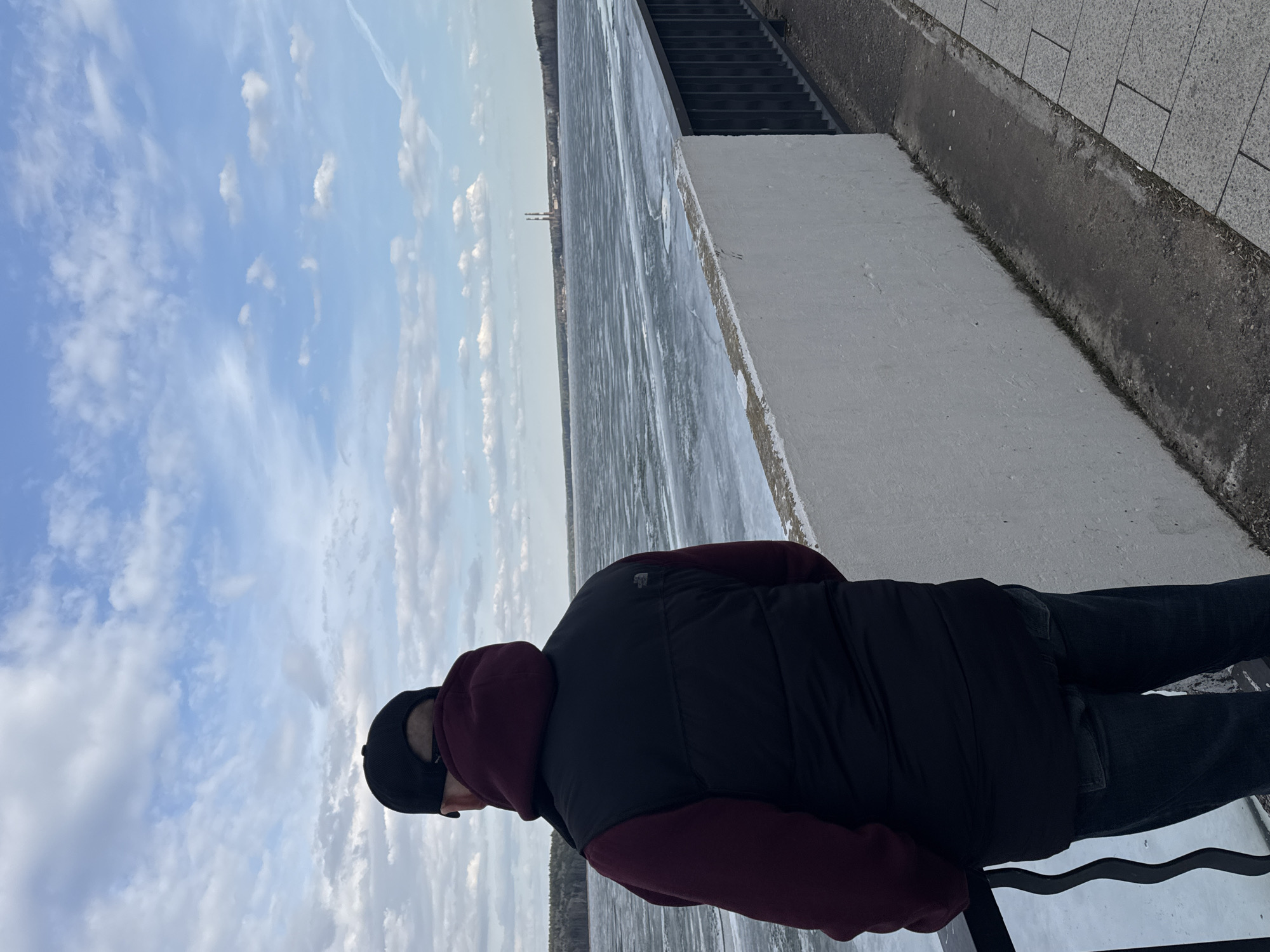 A daytime photograph captures a person standing on an embankment, gazing out over a vast, ice-covered body of water. The person, seen from a slightly elevated rear-left angle, wears a black baseball cap, a burgundy hooded sweatshirt, and a black puffer vest, which has a small white logo (possibly "The North Face") on the back. They are also wearing dark pants.
To their right, a concrete embankment or pier with a grey paved top surface runs parallel to the water. A set of dark steps descends along this structure further into the background. The expansive waterway, likely a river or reservoir, is completely frozen, displaying streaks and patches of ice stretching into the distant horizon.
In the far distance, across the icy expanse, industrial structures or tall chimneys are faintly visible against the sky. The sky above is mostly blue with scattered white clouds, some wispy, some more defined. The overall scene suggests a cold, clear day at a location beside a large, frozen water body. The context of Chaikovsky, Russia, aligns with such winter conditions. The person's posture suggests observation or contemplation of the winter landscape.