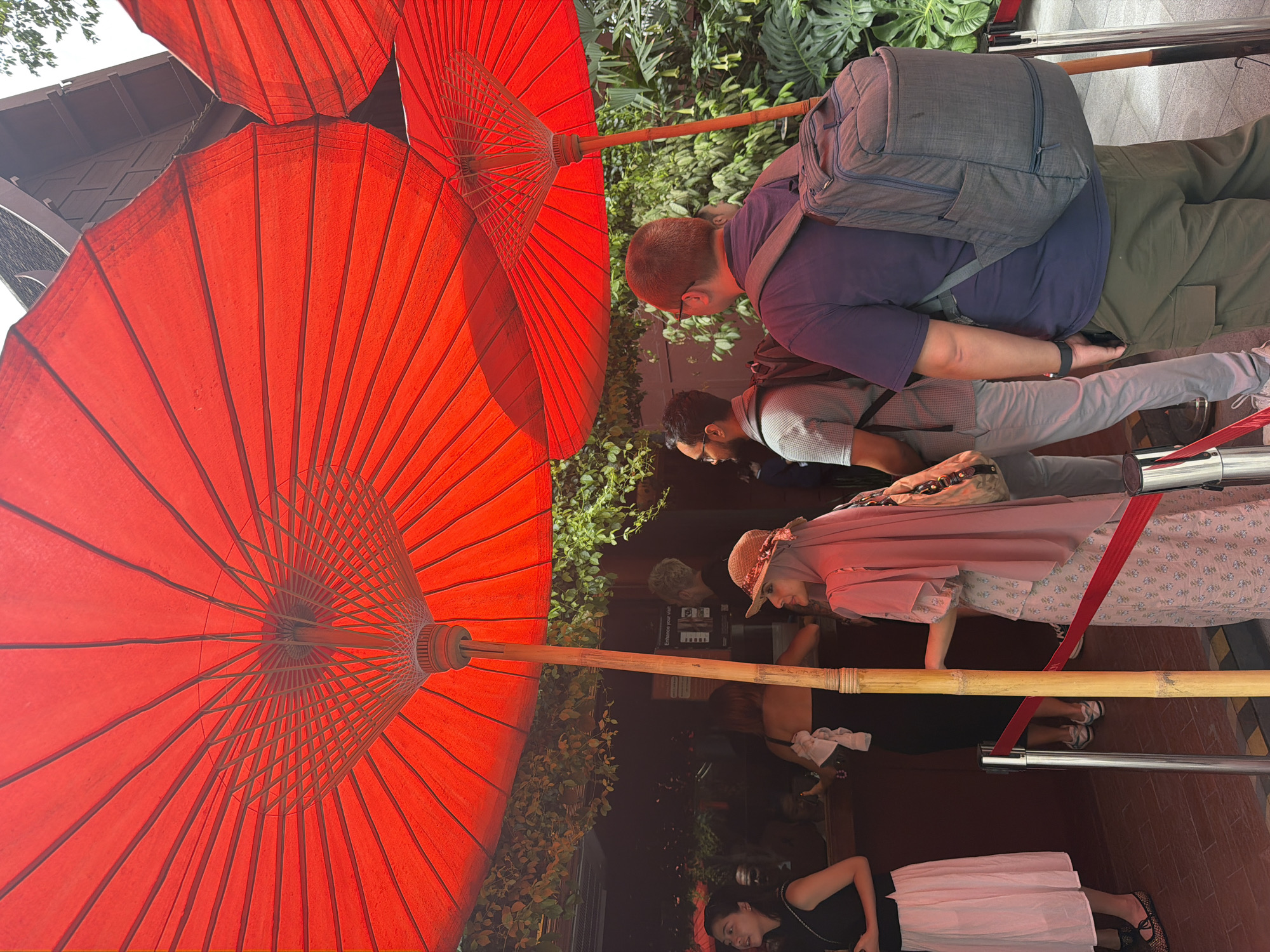 The image depicts a group of people standing in line under large red umbrellas. The setting appears to be an outdoor establishment, possibly a restaurant or a tourist attraction, in Bangkok, Thailand. The architecture visible in the background suggests a traditional Thai style with wooden elements and a sloped roof.
In the foreground, a man with a backpack is turned away from the camera, facing towards the right. To his left, another man with glasses and a backpack is also facing away. In the middle ground, a woman wearing a pink hijab and a patterned dress stands in line. To her left, a woman in a black top and a light pink skirt is visible. Further left, more people are seen inside the establishment, obscured by the umbrellas and the indoor lighting.
The red umbrellas are a prominent feature, providing shade and creating a vibrant visual element. Bamboo poles support the umbrellas, adding to the natural and possibly rustic aesthetic. A red velvet rope barrier indicates a queue. Lush green foliage is visible behind the umbrellas, suggesting a tropical environment common in Bangkok. The overall atmosphere appears relaxed, with people waiting patiently.