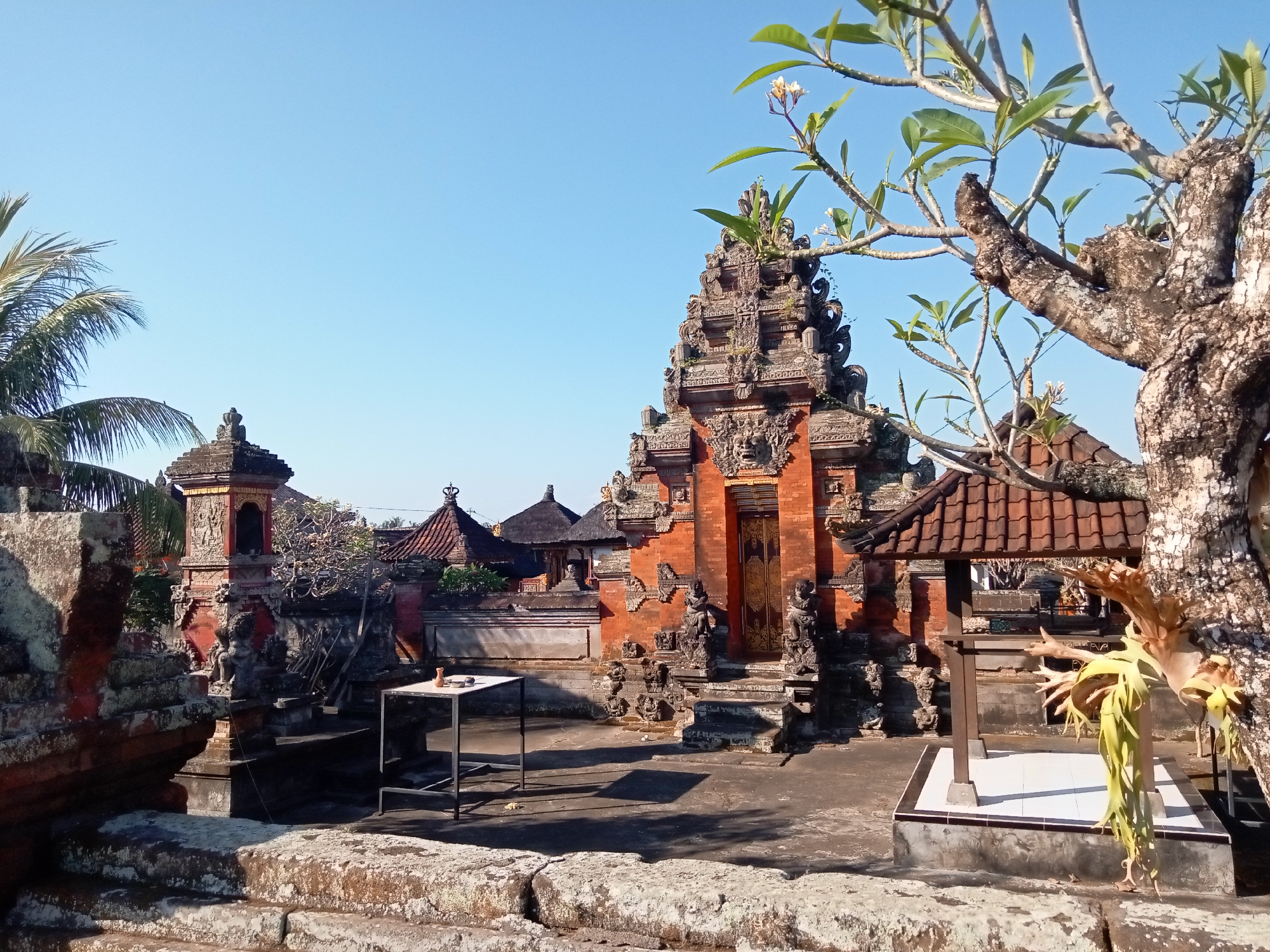 This is a wide shot of a Hindu temple complex under a clear blue sky. The foreground features large, rough-hewn stone steps covered in lichen. In the midground, an ornate red brick temple gate with intricate carvings is visible, topped with a tiered roof structure. Statues flank the entrance, and other temple structures with tiled roofs are scattered throughout the complex. To the left of the main gate, a small, metal table with a white top and a single small object on it is positioned on a paved area. A bare tree trunk with some leaves and a fern-like plant grows in the right midground, partially obscuring a covered structure. The background consists of more temple buildings and a tall palm tree on the far left. The overall impression is one of ancient architecture bathed in bright sunlight.
