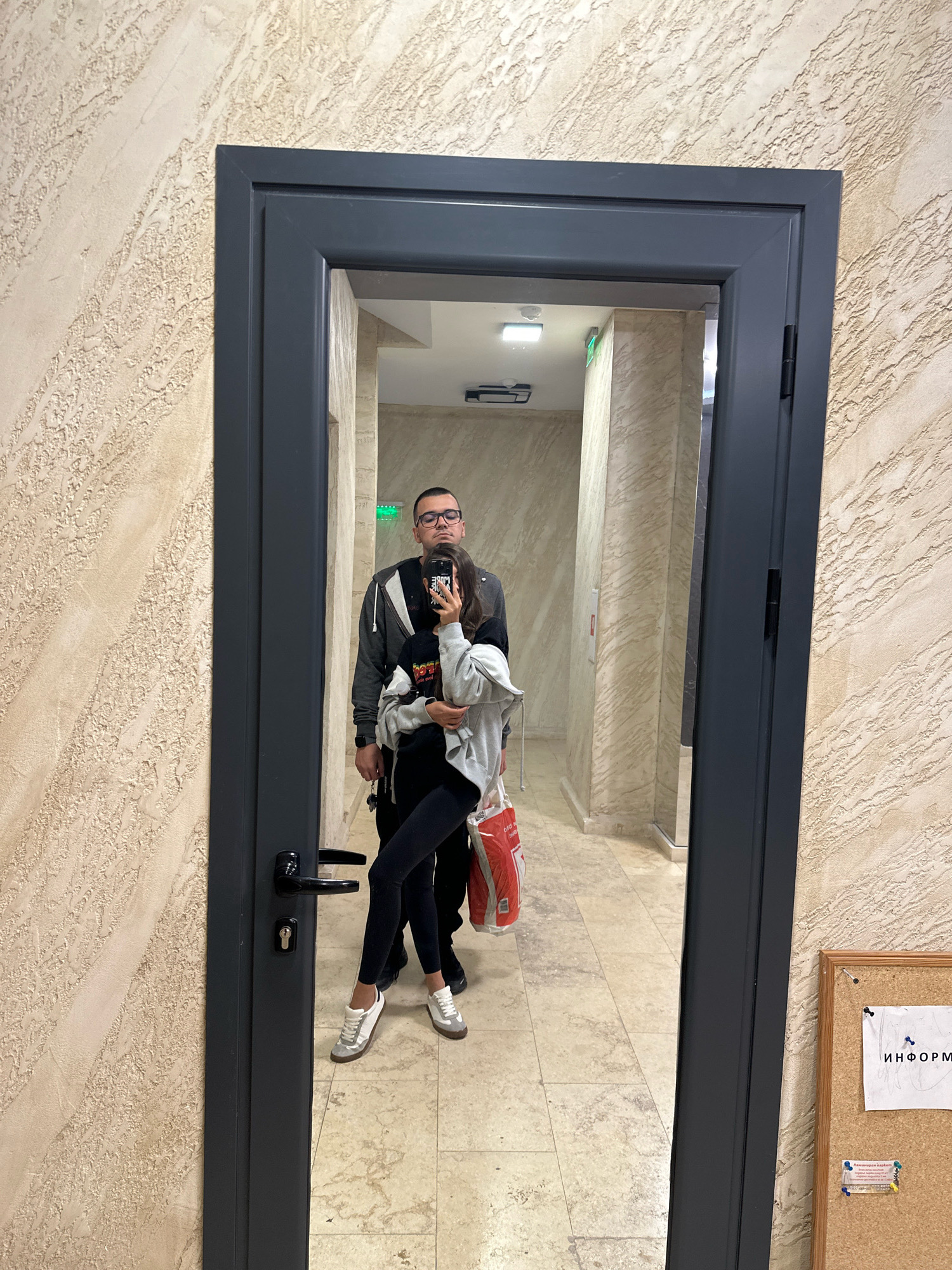 A couple is taking a mirror selfie in what appears to be a modern apartment building hallway. The man, wearing glasses, a grey hoodie, and a dark t-shirt, stands behind the woman. The woman, holding a phone to take the picture, is wearing a grey hoodie over a black t-shirt and black leggings, with white sneakers. She is embracing the man. They are standing on a tiled floor in front of a dark grey door with a silver handle and lock. The hallway walls have a textured beige finish. In the background, a corkboard with a paper notice is visible on the right side. The overall scene suggests a domestic or residential setting, likely in Sofia, Bulgaria, given the presence of Cyrillic text on the corkboard.