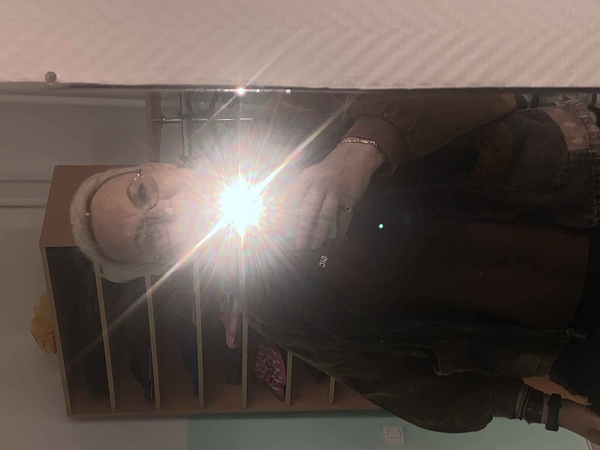 The image shows a person reflected in a mirror, with a strong light source obscuring some details. The person appears to be older, with white hair and glasses. They are wearing a brown jacket and have bracelets on their wrist. A shelf with compartments is visible behind the person in the reflection. The wall behind the shelf is light blue. No information about the city of Chaikovsky, Russia is apparent in the image.