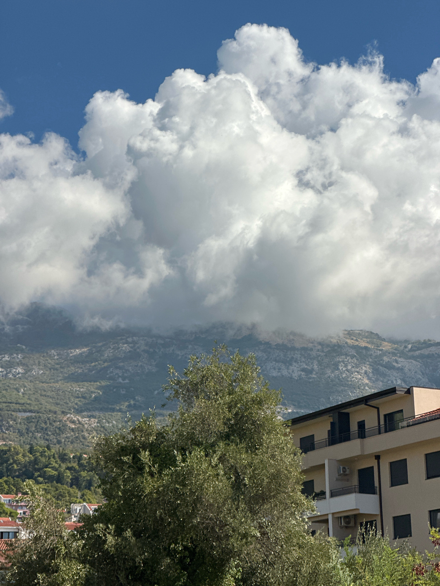 The image depicts a scenic view of the Budva, Montenegro area, characterized by a dramatic sky and mountainous landscape. In the foreground, a modern beige apartment building with dark-framed windows and balconies is partially obscured by lush green trees, likely olive trees given the Mediterranean vegetation. The building appears to be multi-story, with several floors visible.
Behind the building and trees, a vast mountain range stretches across the horizon. The mountains are rugged and covered in sparse vegetation, with some areas appearing rocky and others green. A thick band of white, fluffy clouds hangs low over the mountains, partially obscuring their peaks and giving the impression of a slightly overcast or humid day. Above these clouds, a clear blue sky is visible, dotted with some scattered white clouds. The overall impression is one of a coastal mountainous region bathed in sunlight, with a mix of natural beauty and man-made structures. The image is taken from a slightly elevated perspective, looking towards the mountains.