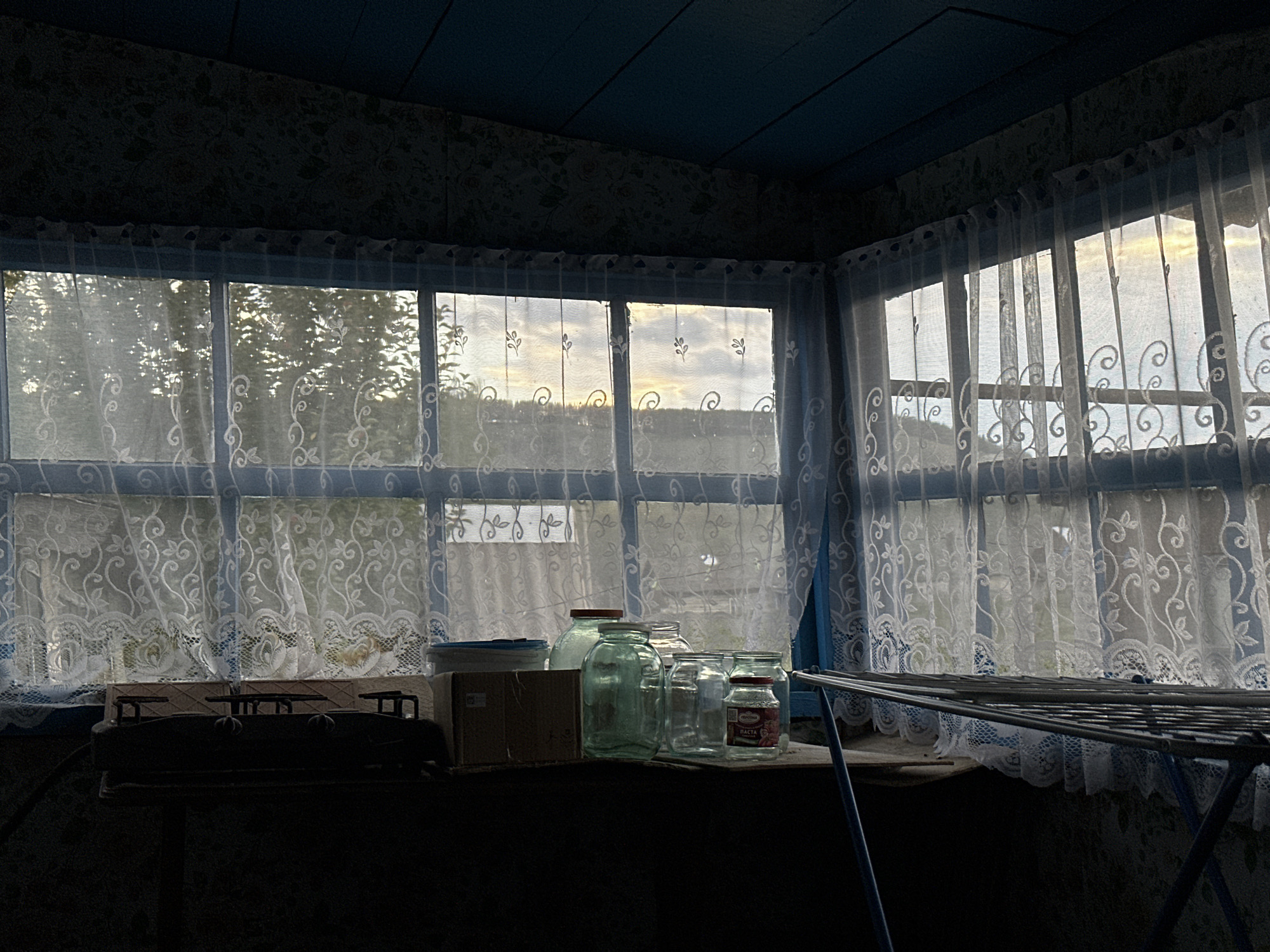 The image depicts the interior of a room with a window overlooking a landscape. The room has blue painted wooden ceiling planks. The walls are covered with patterned wallpaper. A large window with a blue frame is covered by white lace curtains with floral motifs. Through the curtains, trees and a cloudy sky at sunset or sunrise are visible. On a windowsill or shelf in front of the window, there are several glass jars of various sizes, some filled with green liquid or other contents, and a small cardboard box. To the left, a dark object that resembles a scale is partially visible. To the right, a metal clothes drying rack with blue legs is positioned. The overall scene suggests a rustic or domestic setting, possibly a dacha or a simple dwelling in a rural area. The location is identified as Beryozovka, Russia.