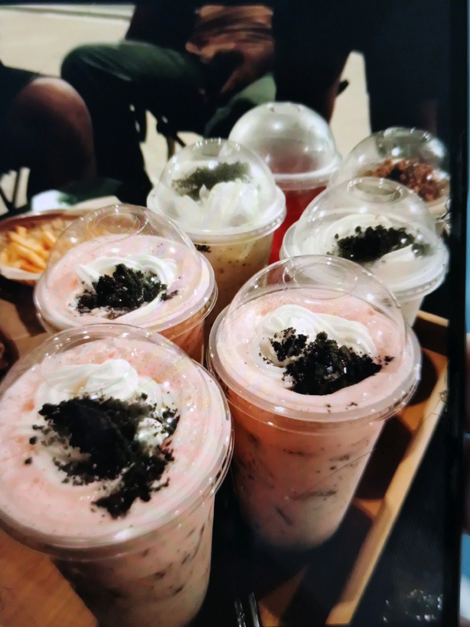 The image shows a close-up view of several plastic cups filled with drinks topped with whipped cream and crushed cookies, likely a dessert or beverage common in cafes. In the background, out of focus, are parts of people, suggesting a social gathering or dining setting. The overall impression is of a casual, possibly urban environment where food and drinks are being served. The detail about the specific location in Caloocan, Philippines, cannot be determined from the visual information provided in the image itself.