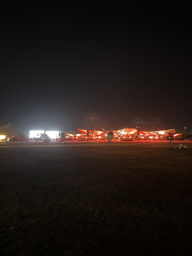 The image shows an outdoor area at night in Indio, United States. The foreground is a dark field or grassy area. In the background, there are structures illuminated with bright lights. To the left, there's a large, brightly lit building or structure. To the right, several structures are lit with orange and red lights. A few vehicles are visible near the illuminated structures. The overall scene is dark, with the only light coming from the structures and some faint light above.