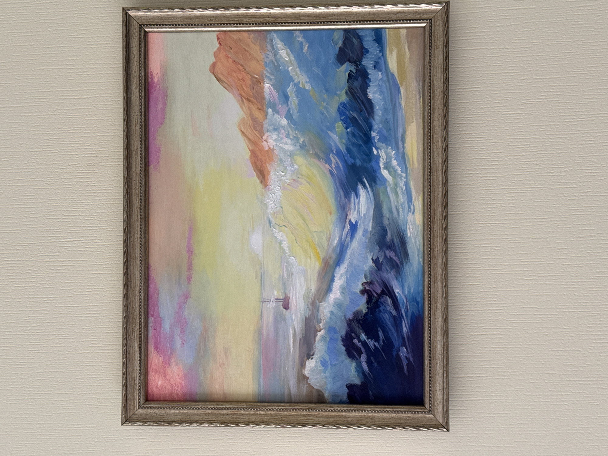 A framed painting depicting a vibrant seascape is centrally displayed against a textured, light-colored wall. The painting, rendered in an impressionistic or expressionistic style with visible brushstrokes, captures large, powerful blue waves crashing, possibly against a cliff face or shore visible on the left, rendered in shades of orange and red. White foam crests the waves, adding to the dynamic motion of the scene. In the calmer waters further out, a small, solitary sailboat with a distinct mast can be seen. The sky or background behind the waves and cliff features a gradient of soft yellow, pink, and purple hues, suggesting either dawn or dusk.
The painting is encased in an ornate, silver-colored frame with a decorative twisted or braided pattern along its inner and outer edges. The wall behind the painting is an off-white or light beige, featuring a subtle horizontal linear texture, likely wallpaper. The scene is well-lit, providing clear visibility of the artwork and its surroundings. There is no visible text in the image. This scene is located in Saint Petersburg, Russia.