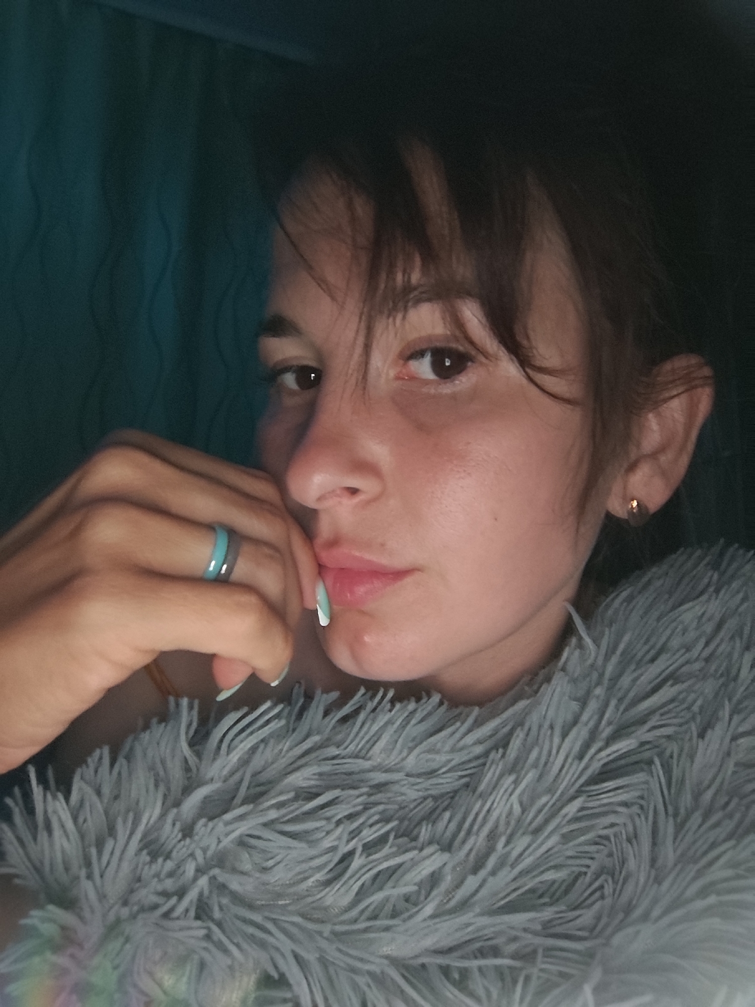 A young woman is pictured indoors, in what appears to be low light or at night. She is partially covered with a fluffy, gray blanket. Her hand, adorned with a ring, is close to her face. She has brown hair and a fair complexion. An earring is visible in her ear. A dark curtain is slightly visible in the background. There is no information to determine the location is in the city of Фоки, Russia.