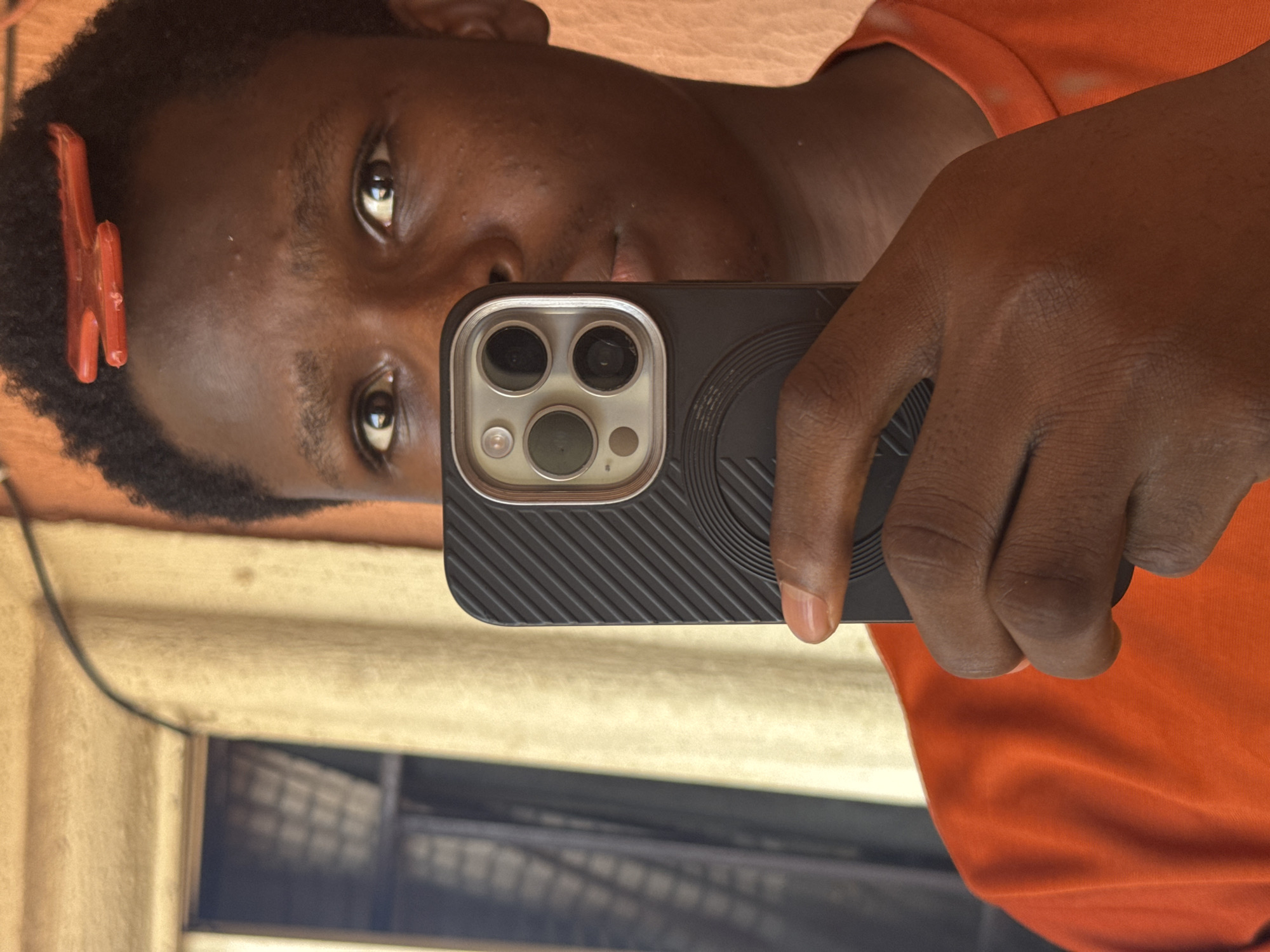 This close-up image, oriented sideways, captures a mirror selfie taken by a person with dark skin. The individual is holding a black smartphone, identifiable by its triple camera array, flash, and LiDAR sensor, housed in a textured protective case.
The person has short, dark, curly hair with a red, M-shaped hair clip positioned on the left side of their head. They are wearing a bright orange t-shirt, parts of which are visible in the reflection. Their dark eyes are looking directly into the phone's camera, which is capturing their reflection.
The setting appears to be indoors, with the reflection revealing warm-toned, possibly beige or light brown, walls. A dark cable runs across the reflected background. Through what appears to be a window or opening in the reflected background, blurred elements resembling a woven basket or architectural features are vaguely visible. The lighting is soft and even, suggesting either diffused daylight or indoor illumination. There is no visible text in the image. The person's expression is neutral. This scene is contextualized as taking place in Onitsha, Nigeria.