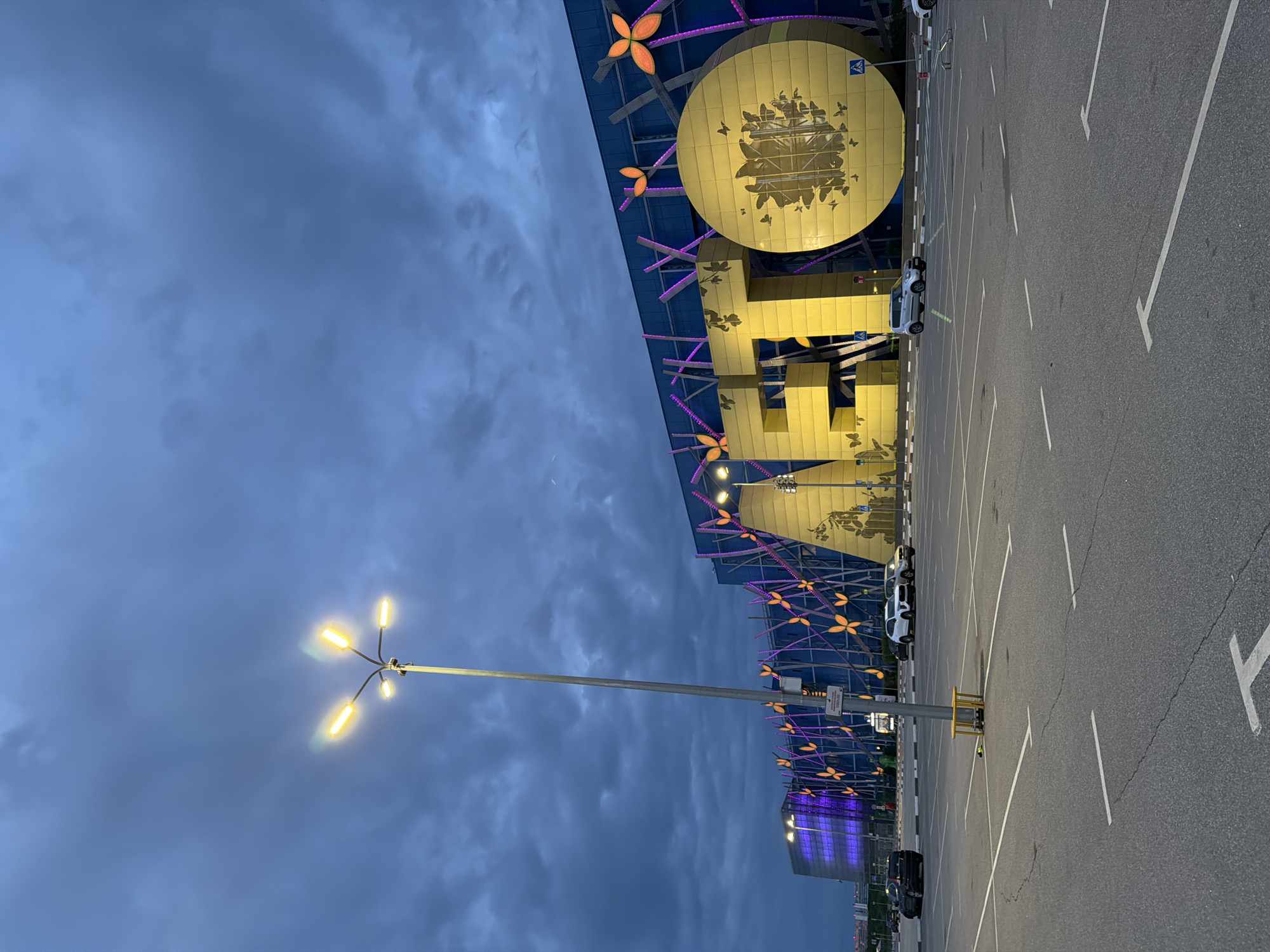 A wide shot captures a large, modern building with a distinctive yellow logo in the city of Unknown, Unknown. The building features a blue facade adorned with decorative elements, including purple vertical lights and orange flower-like accents. The prominent yellow logo appears to be composed of large, blocky letters and a circular shape, possibly spelling out a name or word. The circular element is decorated with numerous small butterfly motifs.
In front of the building is a spacious, mostly empty asphalt parking lot marked with white lines indicating parking spaces. Several cars are parked in the lot, including a black sedan, a white sedan, a silver compact car, and another car further in the background. A tall lamppost with multiple light fixtures stands in the foreground, casting a yellow glow. The sky above is overcast with dark, dramatic clouds, suggesting it might be dusk or a stormy evening. The overall atmosphere is somewhat moody and modern.