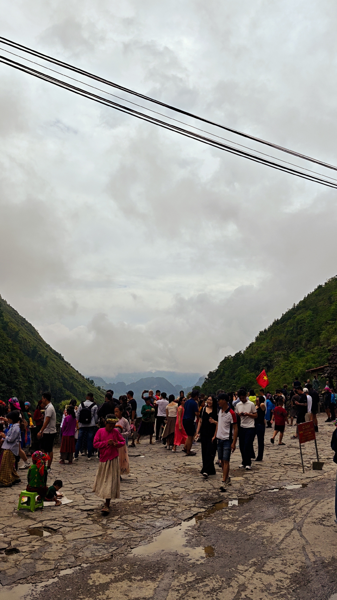 The image depicts a gathering of people on a stone-paved area in Yên Minh Commune, Vietnam, set against a backdrop of mountains under a cloudy sky. A power line is visible at the top of the frame. The crowd is diverse, with individuals of varying ages and attire. Some are taking pictures, while others are walking or standing around. A Vietnamese flag can be seen among the crowd. The ground is uneven and wet, with puddles reflecting the overcast sky. On the left side, someone is sitting on a green stool.