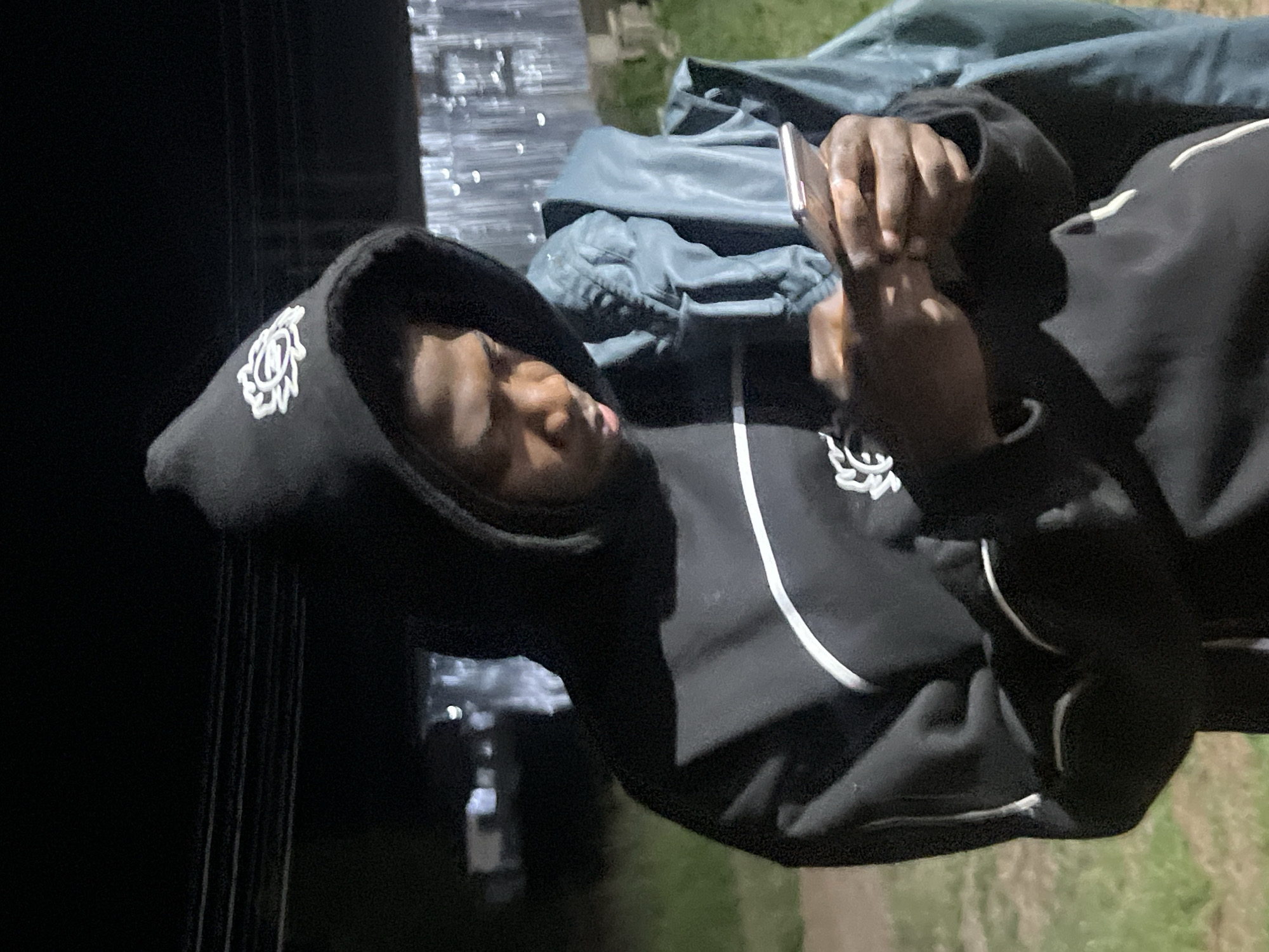 The image captures a close-up of a young Black male wearing a black hooded sweatshirt with a white emblem on the hood and a white stripe across the chest. He is holding a smartphone in his hands and appears to be looking at it, with his head tilted down. He is also wearing a teal-colored jacket draped over his shoulders. The background is dark, suggesting it is nighttime or a dimly lit area, with some out-of-focus lights and what appears to be a corrugated metal structure in the distance. The ground beneath him is covered with grass. The scene is set outdoors, likely in a residential or informal settlement area within Durumi, Nigeria.