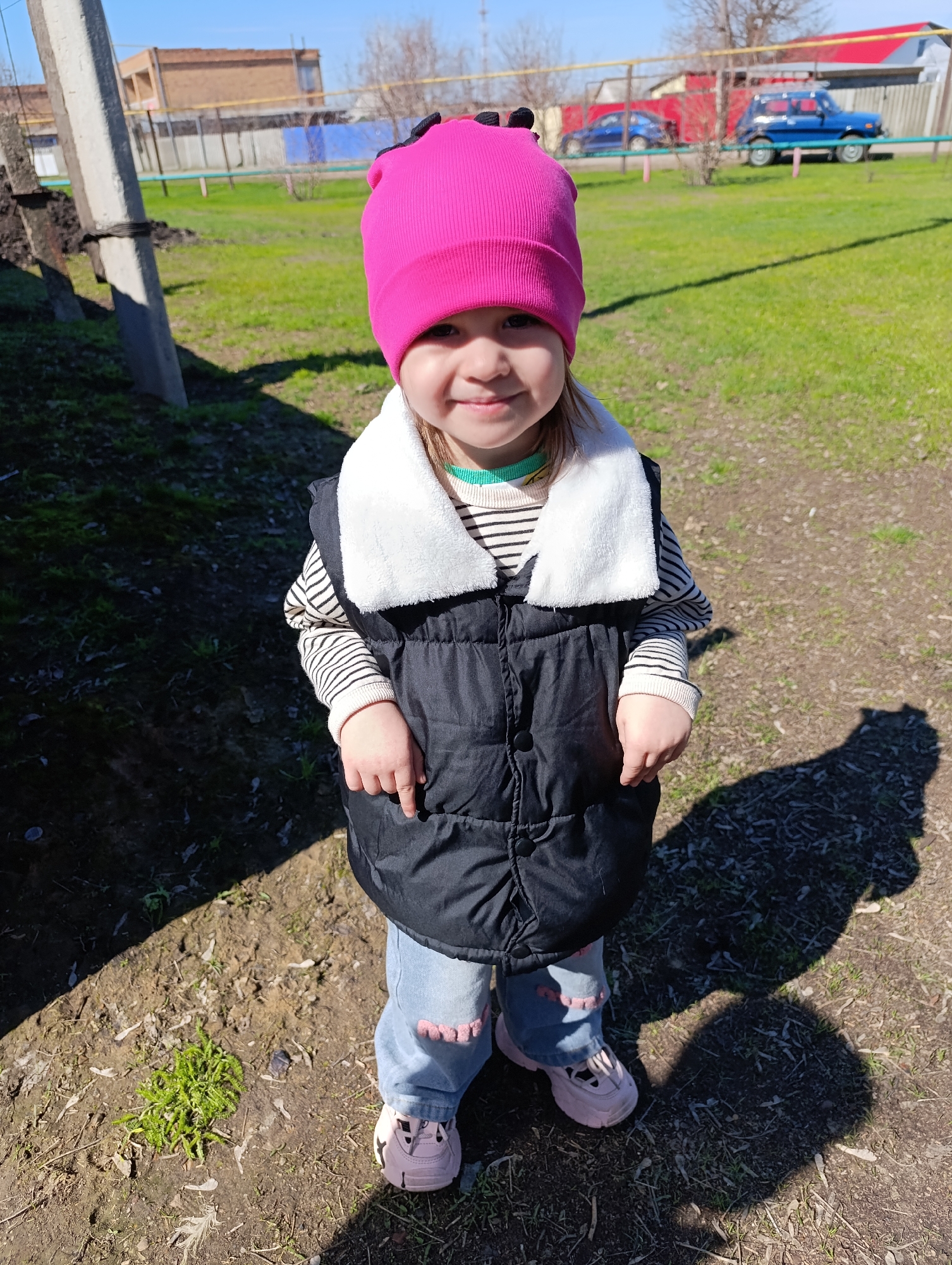 A young child stands on dirt and grass in Уральский, Russia. The child wears a pink hat, a black vest with a white collar over a striped shirt, and blue jeans with pink heart patches on the knees, paired with pink sneakers. The child's shadow falls to the right. In the background, there's a green lawn with scattered leaves, a fence, a blue car, and a small building with a red roof. A utility pole is visible on the left. The weather appears sunny.