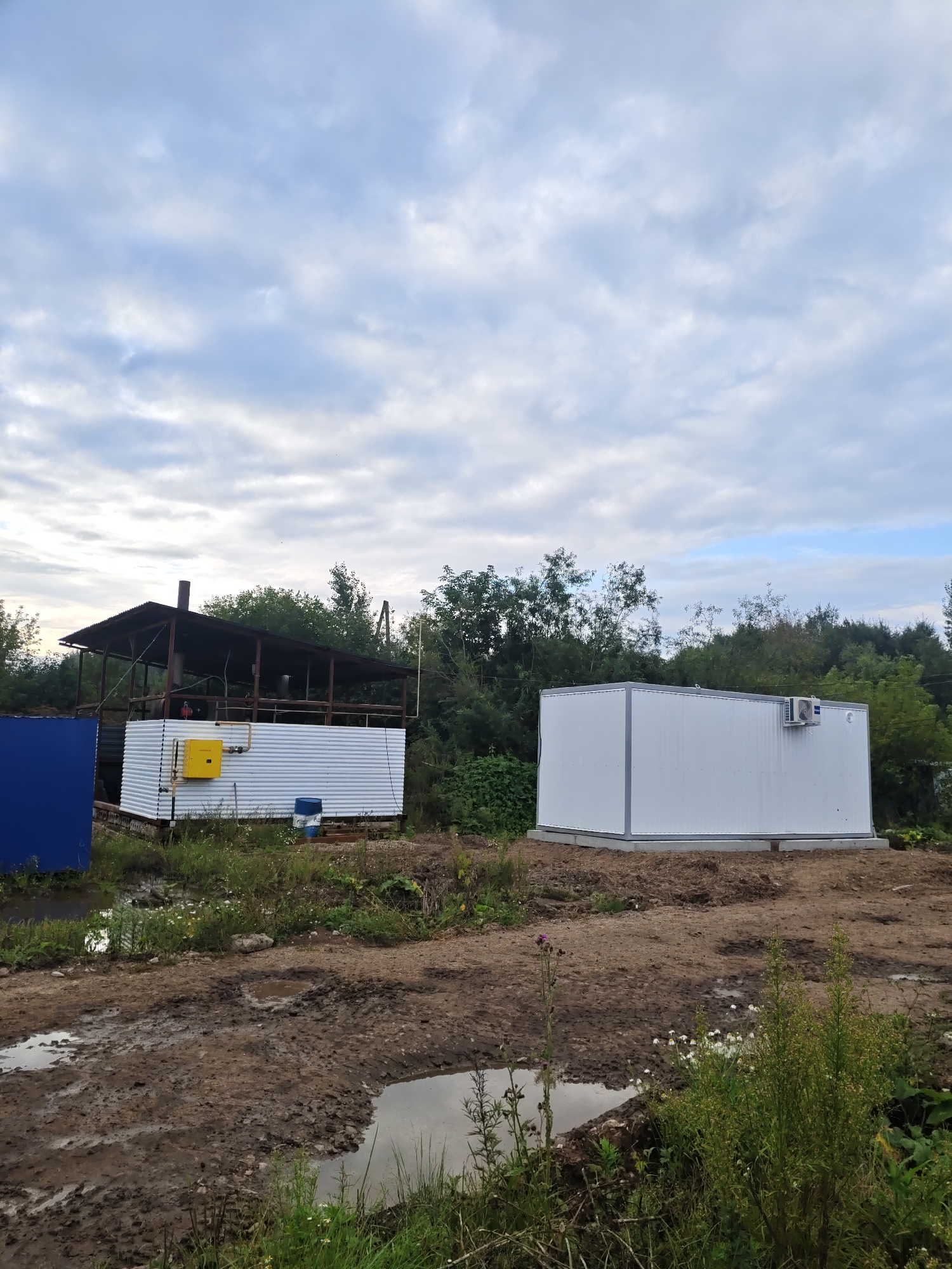 The image depicts an outdoor scene in a semi-developed area. In the foreground, muddy ground with puddles of water is visible, with some sparse vegetation and wildflowers. To the left, a blue metallic structure is partially seen. Behind this, a corrugated metal building with a peaked roof structure supported by metal beams stands. This building has a yellow box mounted on its side, with piping. To the right, a white, rectangular modular building with a grey trim sits on a concrete foundation. An air conditioning unit is mounted on the side of this building. The background is dominated by a line of trees and vegetation, under a cloudy sky. The scene appears to be in Платошино, Russia, suggesting a construction or industrial site. No people are present in the image.