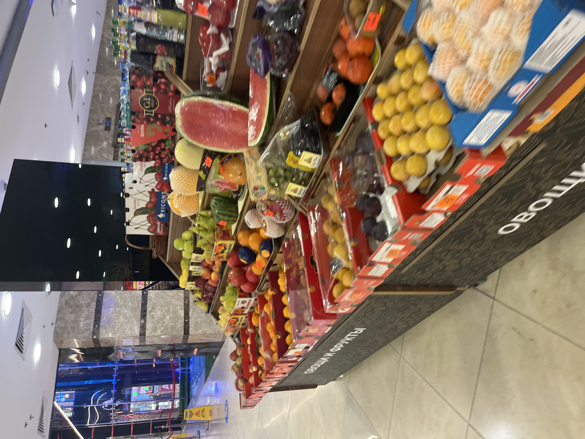 The image shows a produce section in a supermarket, likely in Moscow, Russia. The display features a variety of fruits, including watermelons, apples, pears, oranges, peaches, plums, and grapes. The fruits are arranged on tiered shelves. A sign with Cyrillic text, possibly reading "Овощи и фрукты" (Vegetables and Fruits), is visible on the front of the display. The background includes other supermarket aisles and the store's ceiling and lighting. The overall impression is of a well-stocked, brightly lit retail environment.