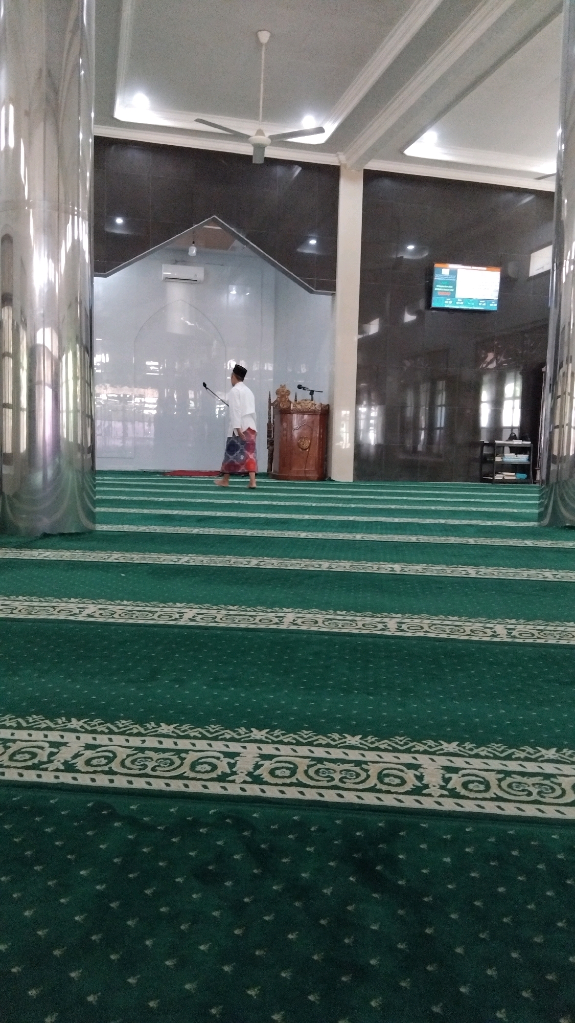 A man wearing a white shirt and a sarong walks across the green carpet of a mosque. He holds a long pole with a cleaning attachment. The mosque has large, shiny pillars and a fan on the ceiling. A digital display shows information on the wall. The image is taken from a low angle, emphasizing the patterns on the carpet. The location is the Baitul Muttaqien Mosque in Batang, Indonesia.