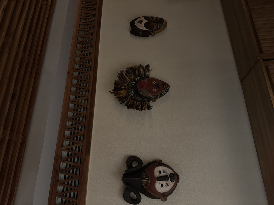 Three tribal masks are mounted on a light-colored wall. The top mask is relatively flat and oval-shaped. The middle mask is rounder and features feathers. The bottom mask has coiled shapes extending from its sides. A decorative wooden panel runs vertically to the left of the masks. To the right, is another panel of wooden slats. The lighting in the image is somewhat dim.