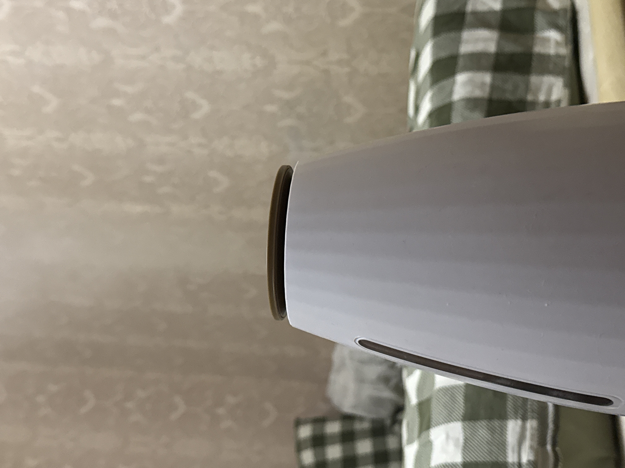 An extreme close-up shows the top of a white humidifier with a brown lid against a blurred background of a beige, patterned wallpaper and bedding with a green and white checkered pattern. The humidifier has a narrow, dark vertical slit on its left side.