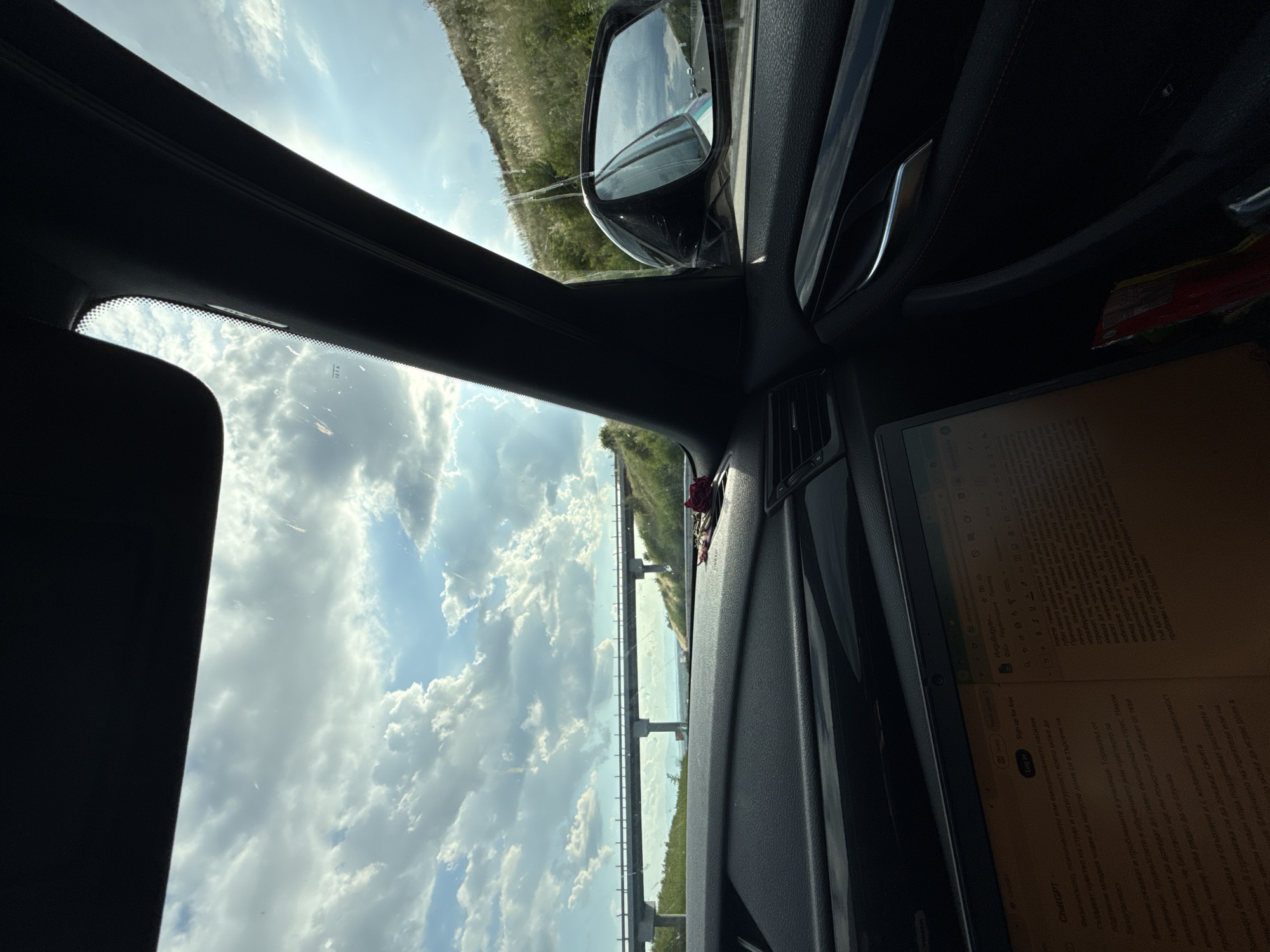 The image captures an interior view of a vehicle's dashboard and windshield, looking out towards a cloudy sky and a distant elevated road or bridge. The foreground is dominated by the dark interior of the car, with a laptop open on the dashboard displaying text. A side-view mirror reflects the road ahead and other vehicles. To the right, a grassy hillside slopes upwards, catching the sunlight. The overall impression is one of travel, with a focus on productivity or work being done while on the move. The specific location of Rupkite, Bulgaria is not visually evident from the provided image.