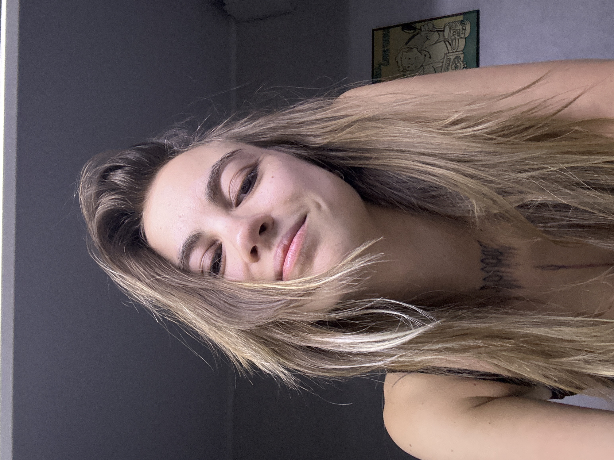A close-up, low-angle, indoor shot shows a young woman with long, blonde, wind-swept hair and brown eyes. She is looking up at the camera and smiling slightly. Her head is tilted to her left, and her hair is falling over her face. She is wearing a black tank top, and a tattoo is visible on her chest and collarbone. A framed picture with cartoon imagery hangs on the wall behind her, to her right. The lighting is dim, and the background is a dark gray wall. The photo appears to have been taken in a private residence in Moscow, Russia.
