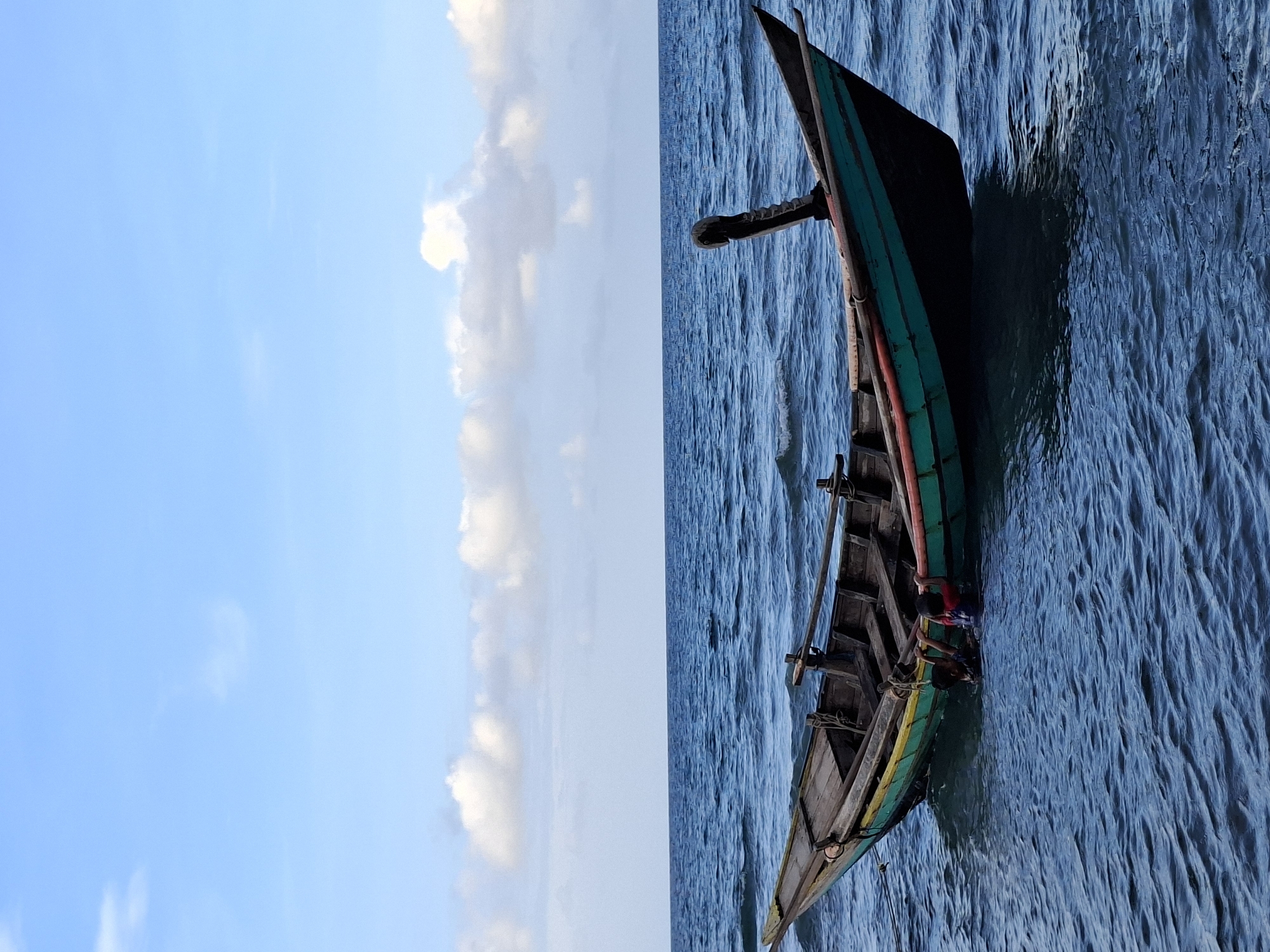 A vertical panoramic shot depicts a boat afloat on the ocean. On the left side of the scene, a clear blue sky is populated with scattered clouds. The right side reveals a wooden boat colored green, yellow, and red. Visible within the boat are figures who appear to be people. The boat is situated on blue waters, which are lightly rippled. It is presumed to be taken in Lhokseumawe, Indonesia.