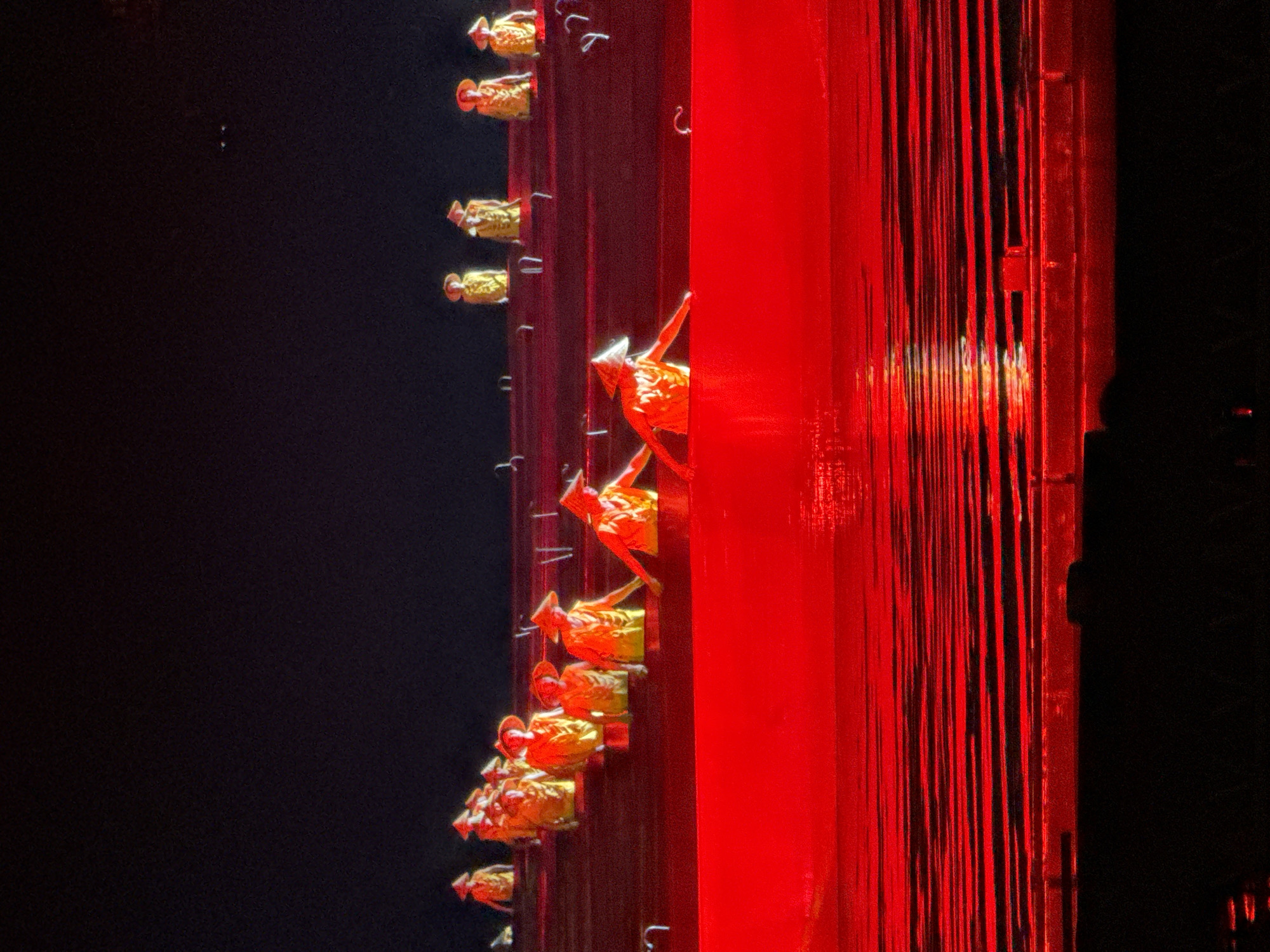 The image shows a vertical arrangement of elements against a black background. On the left, there is a dark void. To the right, a vertical structure is visible, with multiple small, golden figures attached to it. The figures are reminiscent of small human forms, adorned with headwear, and are arranged in a descending line. Adjacent to this, a vibrant red structure, featuring vertical lines or stripes, takes up a substantial portion of the right side of the frame.
Without further context, the specific location cannot be determined. However, based on similar imagery, the scene is likely from a show at the "Dreamlike Lijiang" water show in Guilin, China.