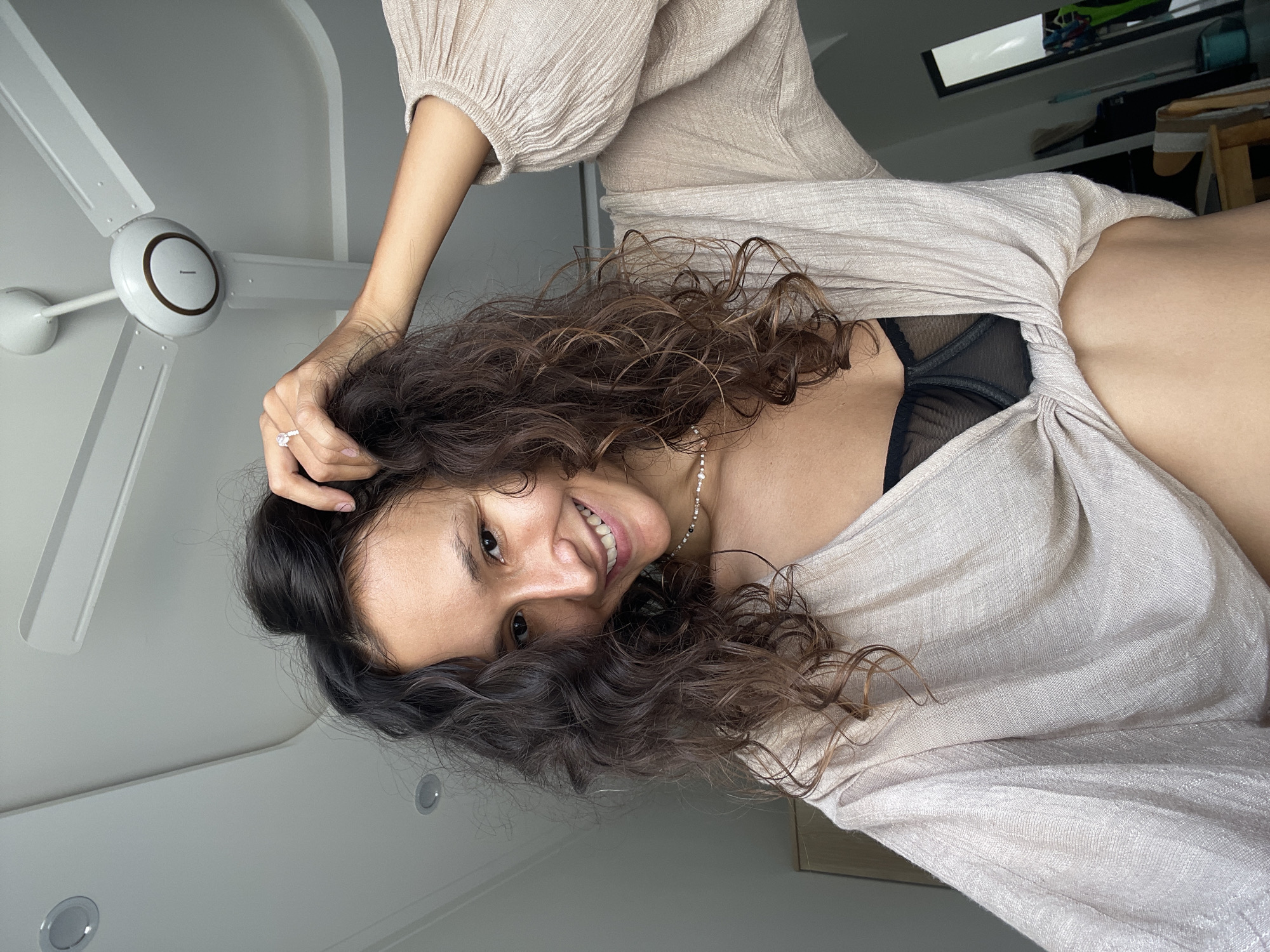 The image, originally rotated 90 degrees counter-clockwise, captures a cheerful young woman looking directly at the camera indoors. She has long, dark, voluminous curly hair and is smiling broadly, showing her teeth. Her head is tilted, and her right hand is gently touching her hair; she wears a prominent light-colored stone ring on her right ring finger. A delicate beaded necklace is visible around her neck.
She is dressed in a light-colored, likely beige or off-white, loose-fitting top with gathered sleeves. A black sheer lace bra is visible beneath the neckline of her top.
The background shows a white ceiling with a modern white ceiling fan, featuring four blades and a central motor housing with a "Panasonic" logo. Recessed lights are also visible on the ceiling. The bright illumination suggests it is daytime. The overall mood is happy and relaxed. Based on the provided context, the image was taken in Mũi Né, Vietnam.