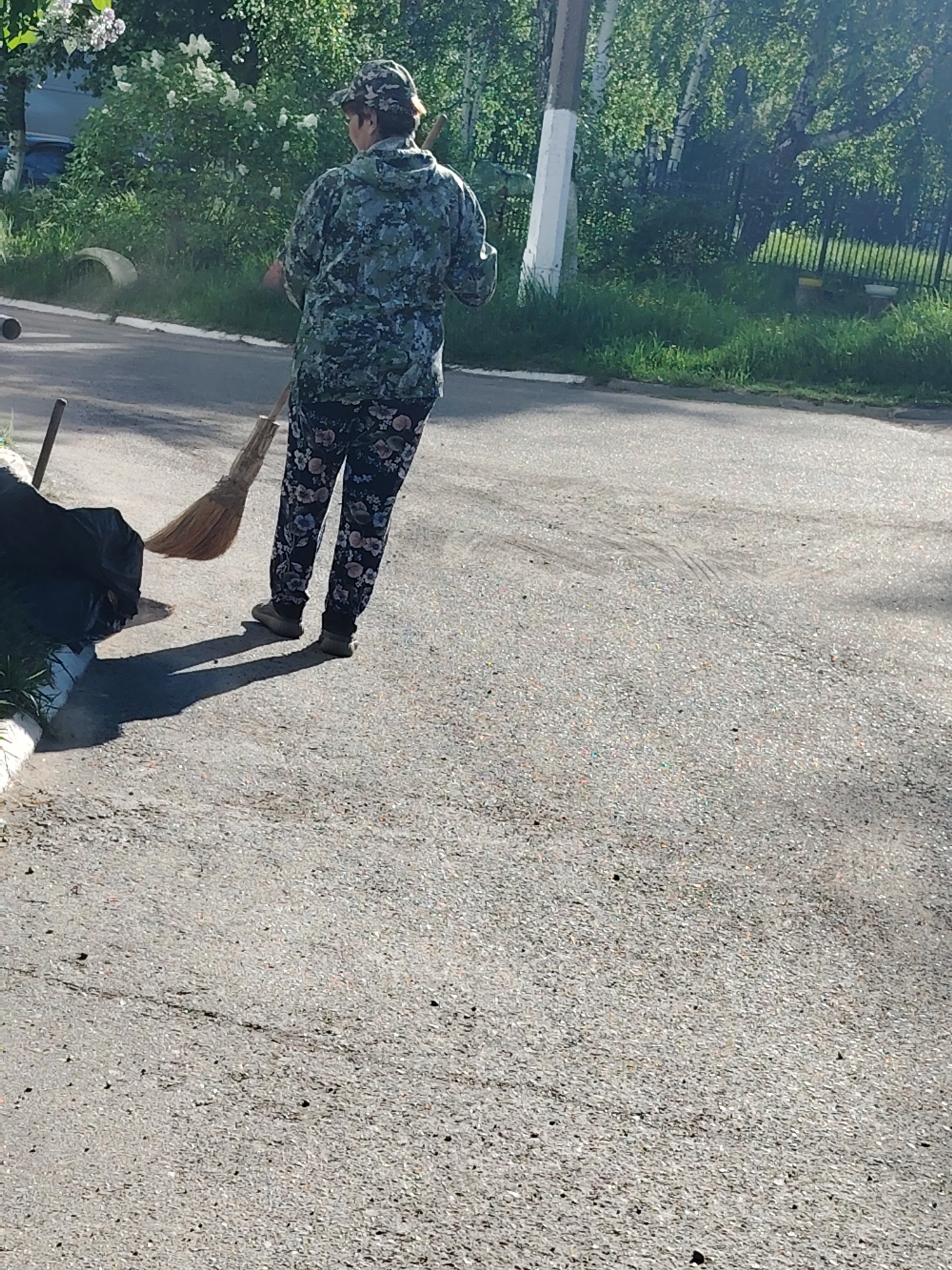A person sweeps a paved street in Chaikovsky, Russia. The individual, viewed from behind, wears a camouflage jacket, floral-patterned pants, a cap, and dark shoes. They hold a broom and sweep debris toward a black trash bag. To the left, a long-handled tool rests near the trash bag. In the background, greenery, including trees and shrubs, is visible, along with a painted pole. The street is light gray, showing signs of wear. Sunlight casts shadows.