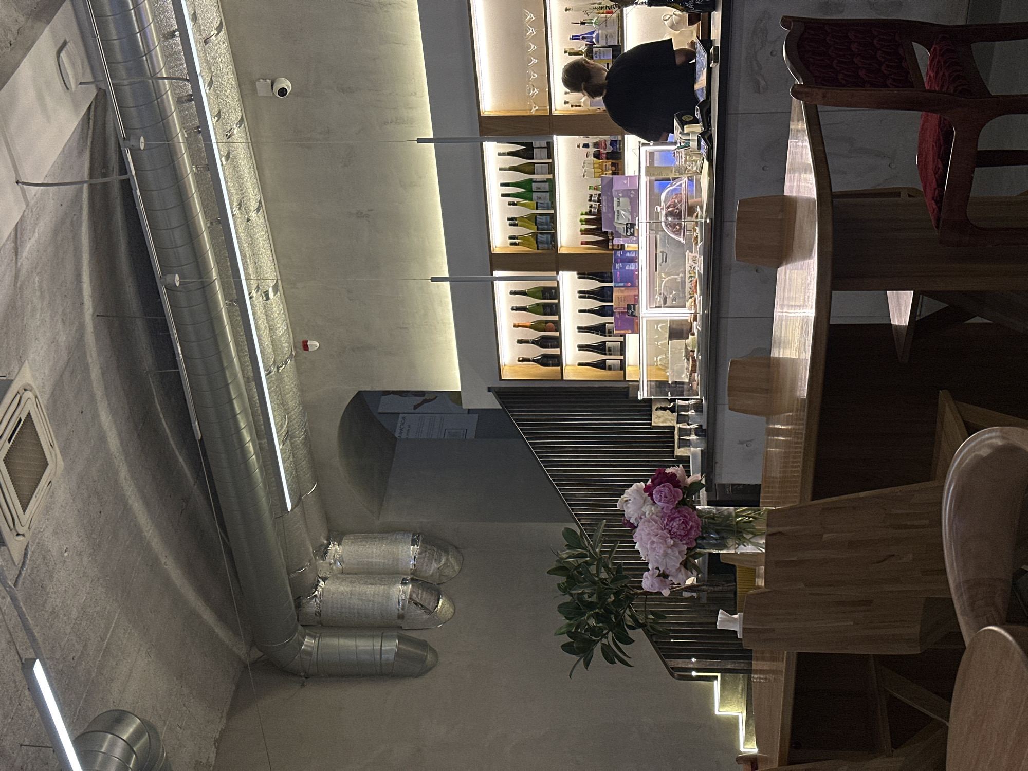 The image shows the interior of a dimly lit establishment, possibly a cafe or bar, with a minimalist, industrial aesthetic. The ceiling is exposed concrete with prominent silver ventilation pipes and modern lighting fixtures. In the foreground, several wooden tables and chairs are visible. To the left, a staircase with illuminated steps leads upwards. A bouquet of pink and purple peonies in a glass vase sits on one of the tables. In the background, a counter displays various bottles of alcohol on shelves illuminated by soft lighting. A person, seen from behind wearing a black shirt, is standing behind the counter, appearing to be a staff member. The overall atmosphere is subdued and contemporary. The specific location in Moscow, Russia cannot be determined from the visual information provided.